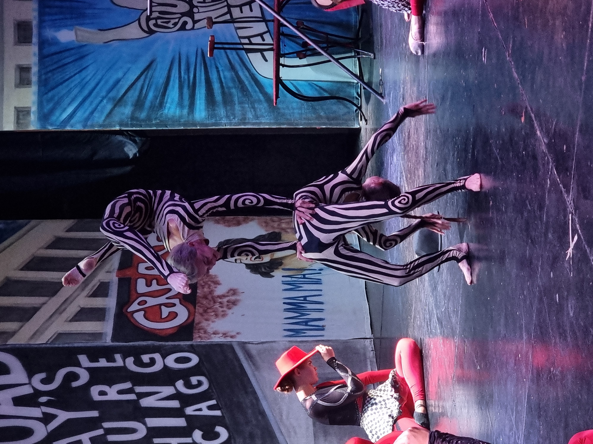 In a theatrical setting, two acrobats in black and white striped costumes are performing a human pyramid. The acrobat at the bottom is on their hands and knees, supporting the weight of the acrobat above. The acrobat on top is balancing in a handstand with their legs extended. A third performer, wearing a red hat and a black and white checkered skirt, is seated on the stage to the left, looking down. Behind the performers are large painted backdrops. One backdrop on the left depicts text that appears to say "ROAD AY'S URE HING CAGO". Another backdrop behind the acrobats features the words "MAMMA MIA!". To the right is a blue backdrop with a graphic of a hand holding a flame and text that reads "STU NIC EVE". A red table with black legs and a black stand are also visible on the right side of the stage. The stage floor is dark and reflective.
