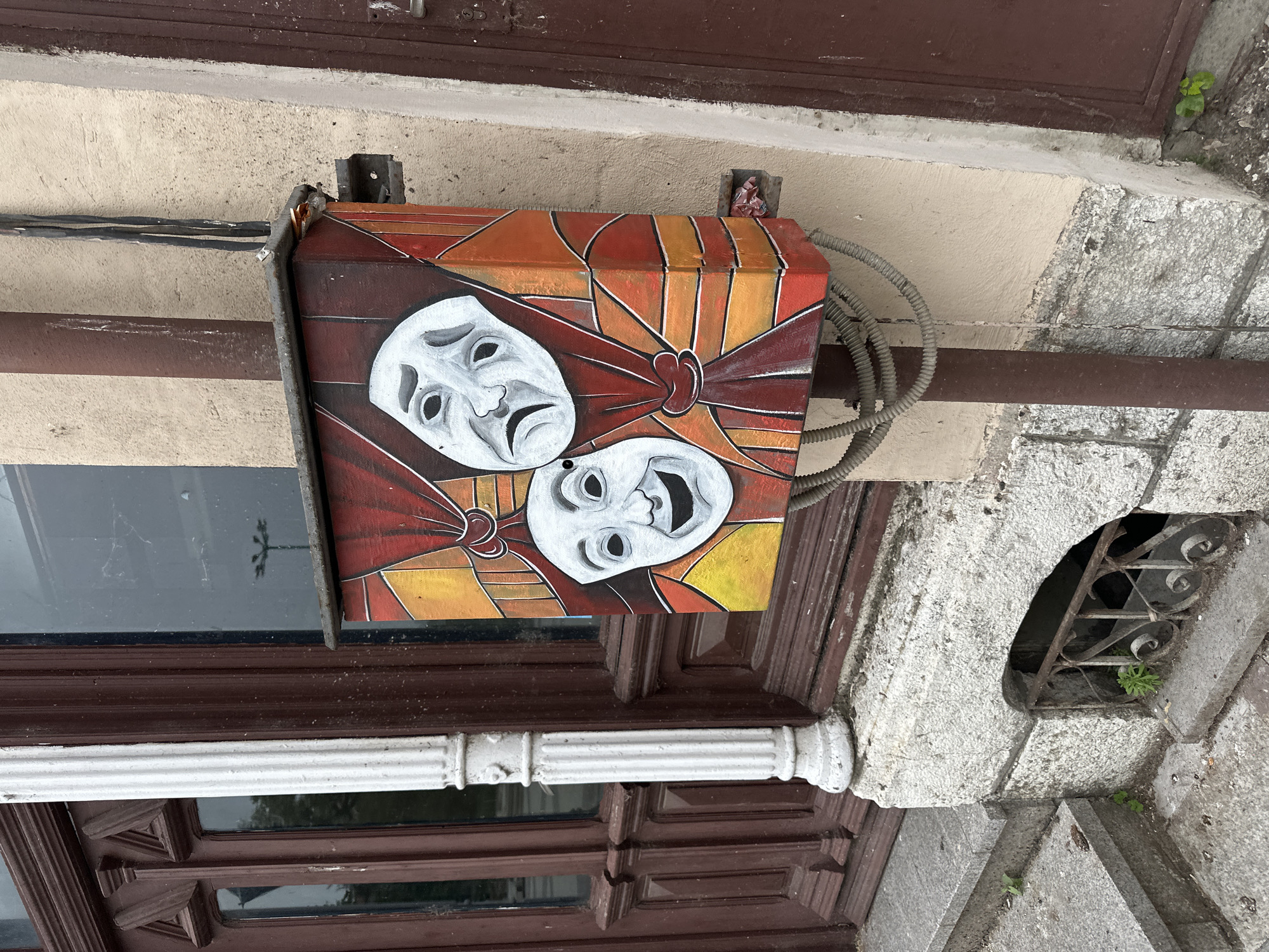 Mounted on the exterior wall of a building in Vidin, Bulgaria, is an electrical box decorated with the iconic theatrical masks of comedy and tragedy. The box is painted with a color scheme of red, orange, and yellow, with the masks painted white with black outlines to accentuate their features. A thick, gray electrical conduit is connected to the box. Beneath the box is a wooden window frame with dark brown panels and a white decorative trim. To the right, the stone facade of the building has a small, gated opening, possibly a basement window.