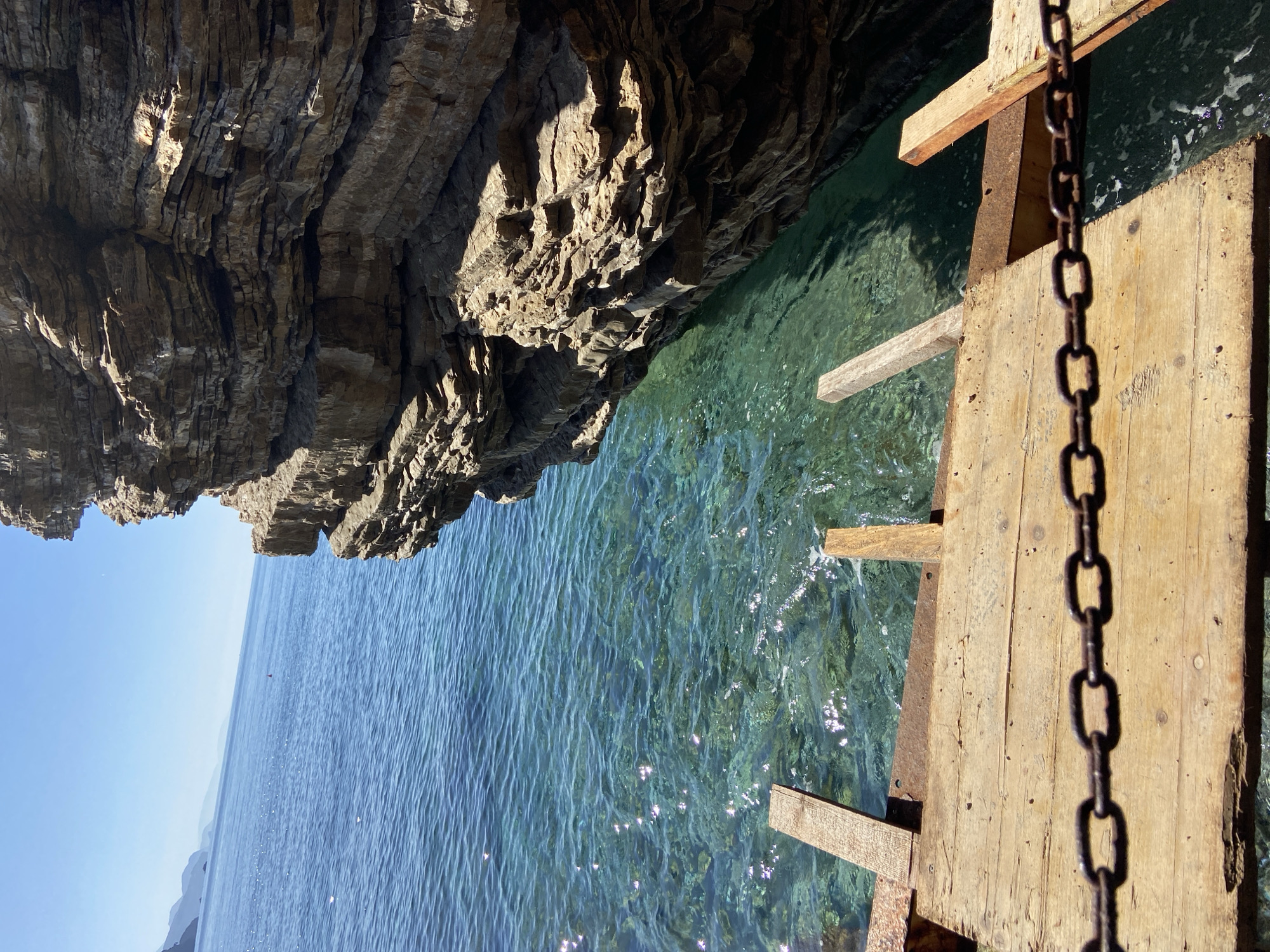 A slightly tilted, eye-level shot shows a rocky overhang and clear blue-green water, with a weathered wooden platform and chain in the foreground. The rocky overhang is on the upper left, with its craggy layers casting shadows. The water below is clear, with light reflecting on its surface. The wooden platform is on the upper right, constructed with planks and support beams. A dark, heavy chain hangs vertically against the wood. The distant horizon suggests an open sea or lake. This scene is possibly a coastal view near Sveti Stefan, Montenegro, judging by the landscape.