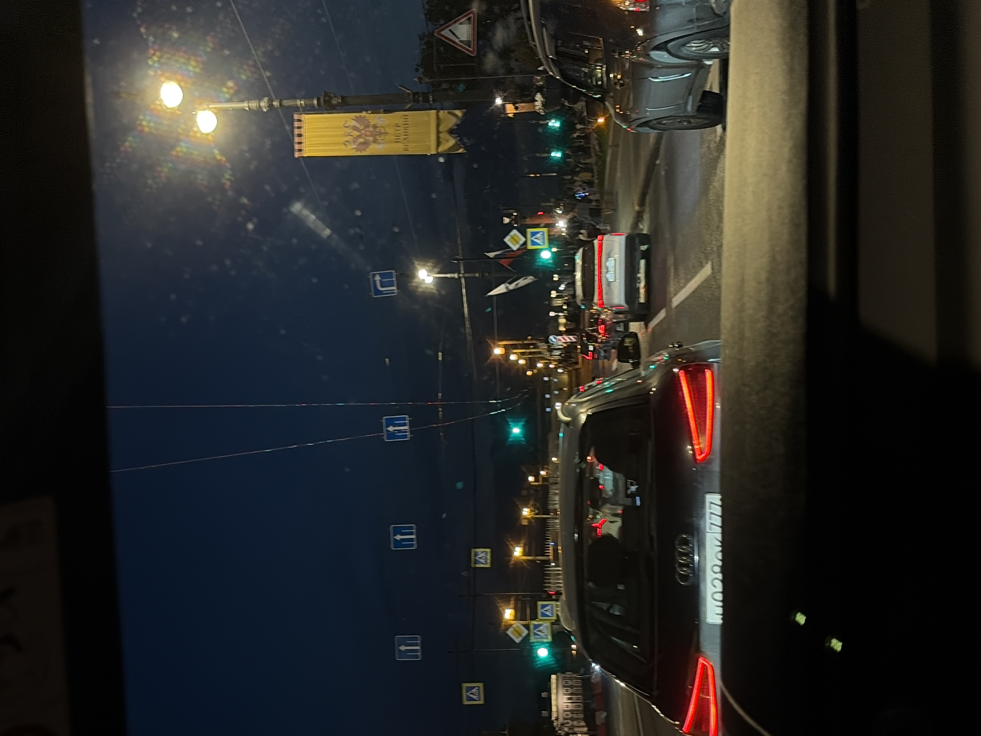The image depicts a nighttime street scene in Saint Petersburg, Russia, as viewed from inside a vehicle. The sky is dark blue, suggesting either late evening or early morning. The street is illuminated by streetlights and the headlights and taillights of vehicles. Several cars are visible, indicating traffic congestion. Road signs, including pedestrian crossing signs and directional signs, are present. A tall banner with a yellow background and a double-headed eagle emblem hangs from a lamppost, a symbol associated with Russia. Flags, possibly the Russian flag, are also visible. The overall impression is of a city at night, with its usual urban activity.