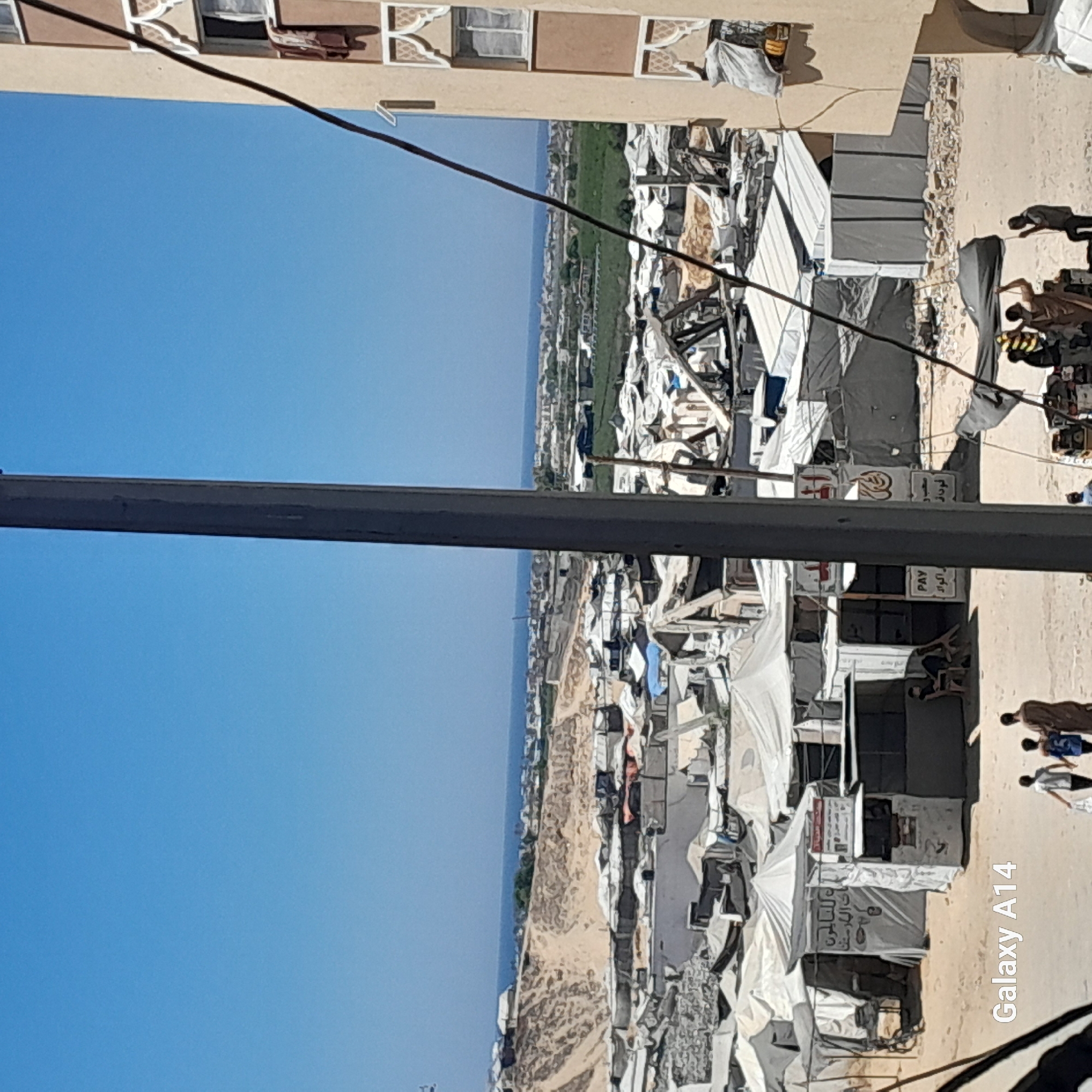 The image depicts a refugee camp in Khan Yunis, Palestinian Territories. The scene is characterized by numerous tents and makeshift shelters set up on sandy terrain. In the foreground, a tall, dark pole runs vertically, obscuring part of the view. To the right, a section of a beige building with decorative window frames is visible. Several people are present in the scene. In the middle ground, a small stand with signage displaying Arabic text and the word "PAY" is set up, with a person sitting on a chair nearby. Further back, more tents and structures are densely packed. Beyond the camp, a body of water (likely the sea) is visible in the distance, with a hazy line of land and buildings on the horizon. The overall impression is one of a densely populated area with basic living conditions. The "Galaxy A14" watermark is present in the lower left corner, indicating the device used to capture the image.