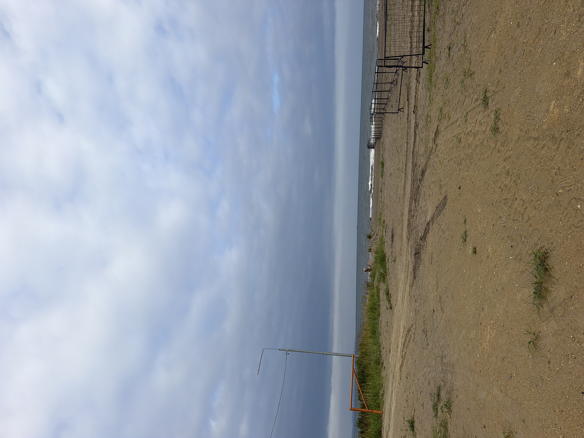 An outdoor, vertical, medium shot captures a beach scene under a cloudy sky. The foreground is dominated by a sandy beach with tire tracks visible. A low, orange gate-like structure stands on the left, adjacent to a patch of green grass and a tall, slender lamp post. Further back, a metal fence stretches along the beach towards the right side of the frame. The sea is visible in the background with moderate waves. The sky is filled with heavy, dark clouds, suggesting overcast weather. The location is identified as Aniva, Russia. No people are visible in the frame.