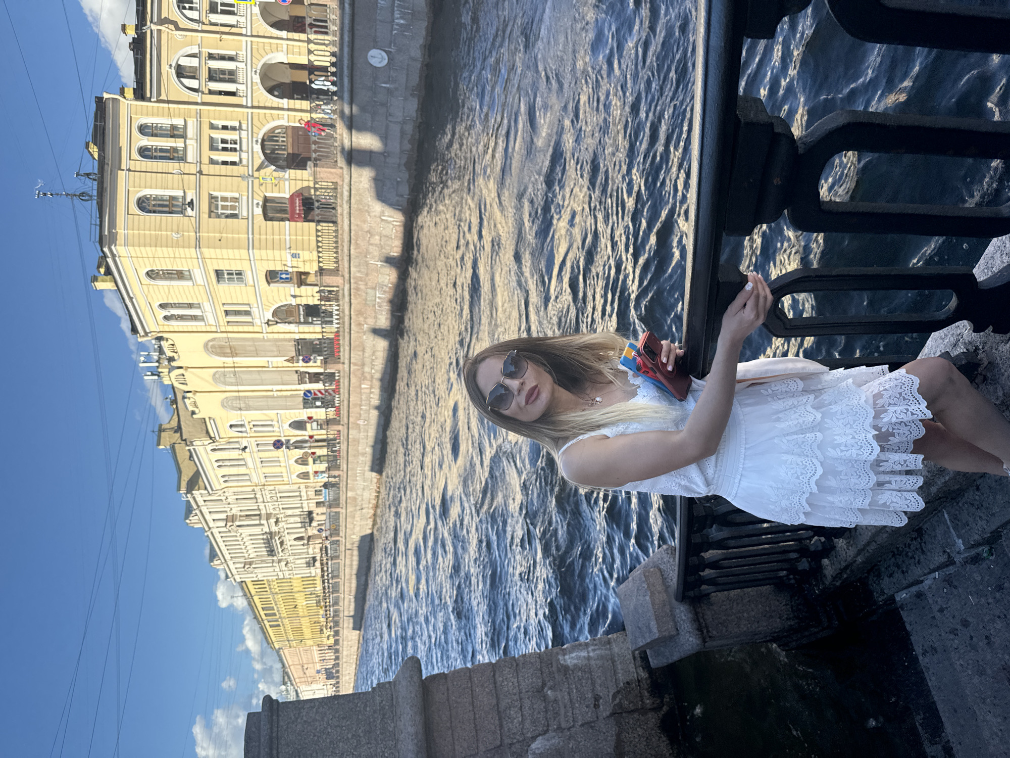 A young woman with blonde hair and sunglasses is sitting on a stone embankment, leaning against a black metal railing. She is wearing a white lace dress and holding a red and blue object in her hand, possibly a phone or a small book. The background features a canal with rippling water and a light yellow building with arched windows and decorative details, characteristic of St. Petersburg architecture. The sky is clear and blue, with some scattered clouds, suggesting it is daytime with pleasant weather. The scene is set in St. Petersburg, Russia, evidenced by the architectural style. The woman appears relaxed and is looking towards the camera with a slight smile, conveying a sense of ease and enjoyment. The overall atmosphere is bright and summery. There is no visible text in the image.