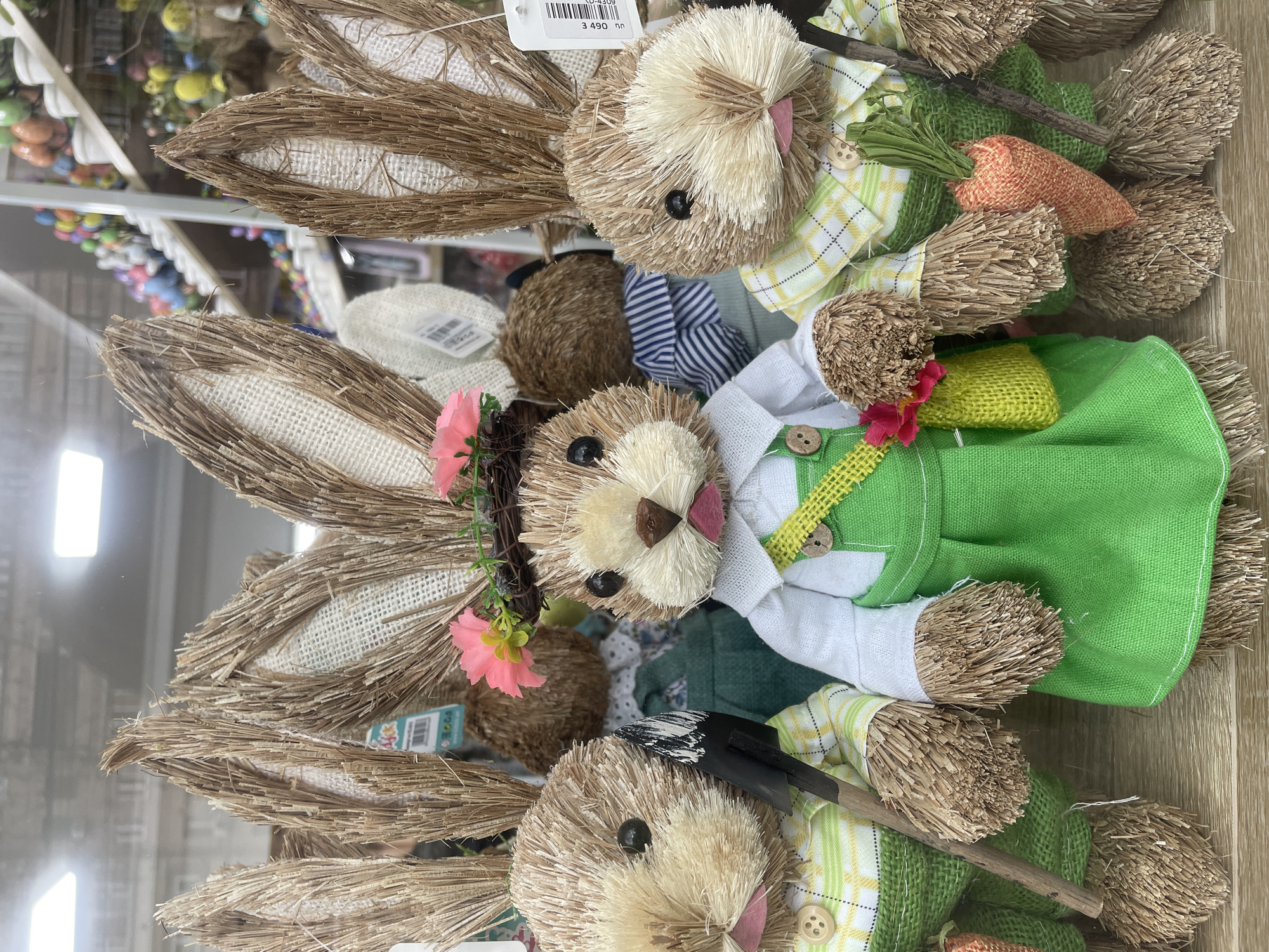 The image shows a collection of handcrafted straw rabbit figurines, likely arranged for sale or display. The location appears to be a store or shop, potentially in Yerevan, Armenia. The rabbits are dressed in various outfits, including green dresses, plaid shirts, and white shirts. Some of the rabbits hold accessories like carrots and gardening tools. The environment is indoors with shelf displays visible in the background. The overall arrangement suggests a spring or Easter theme.