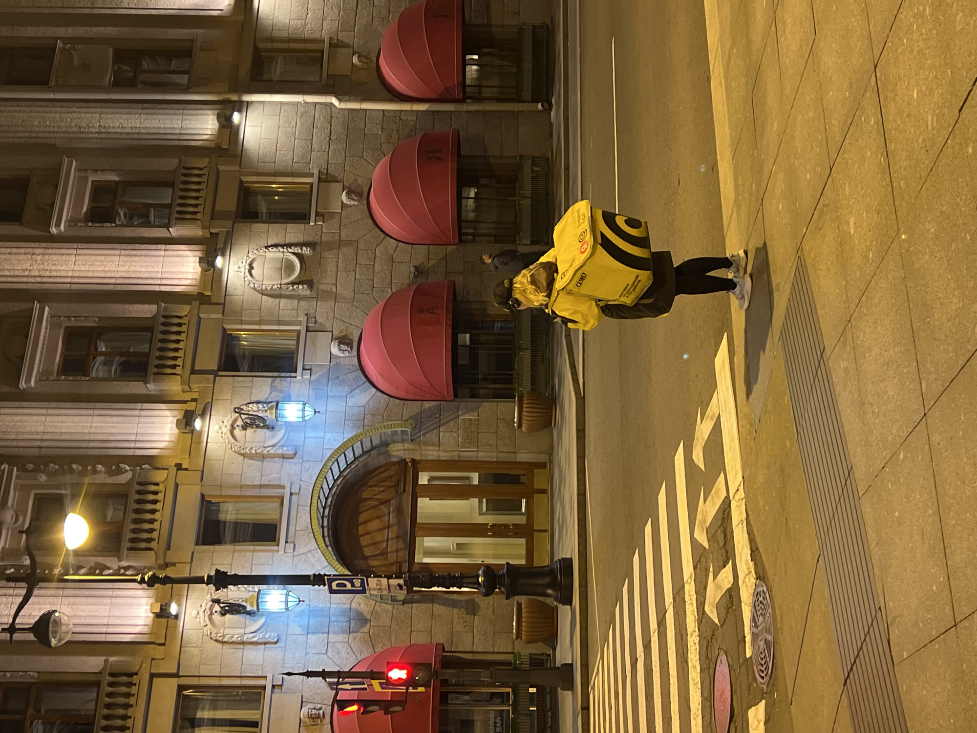 A nighttime street scene shows a woman wearing a yellow delivery backpack standing on a sidewalk next to a street with painted pedestrian crosswalk markings. The architectural facade of a building, possibly a hotel, is visible to the left, featuring awnings and exterior lighting. A streetlamp is also visible. The location is the Grand Hotel Europe in Saint Petersburg, Russia.