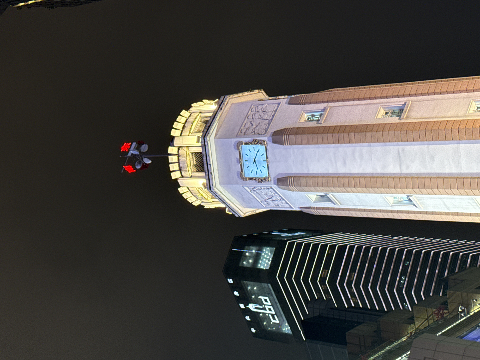 The image captures a night scene in Yuzhong District, Chongqing, China, featuring illuminated buildings. The most prominent is a historic clock tower-style building with a blue clock face, decorated stonework, and small windows. Below it is a modern high-rise with chevron-shaped lighting on its facade. A street light with red blossoms is visible to the side of the clock tower. The background is a dark sky.