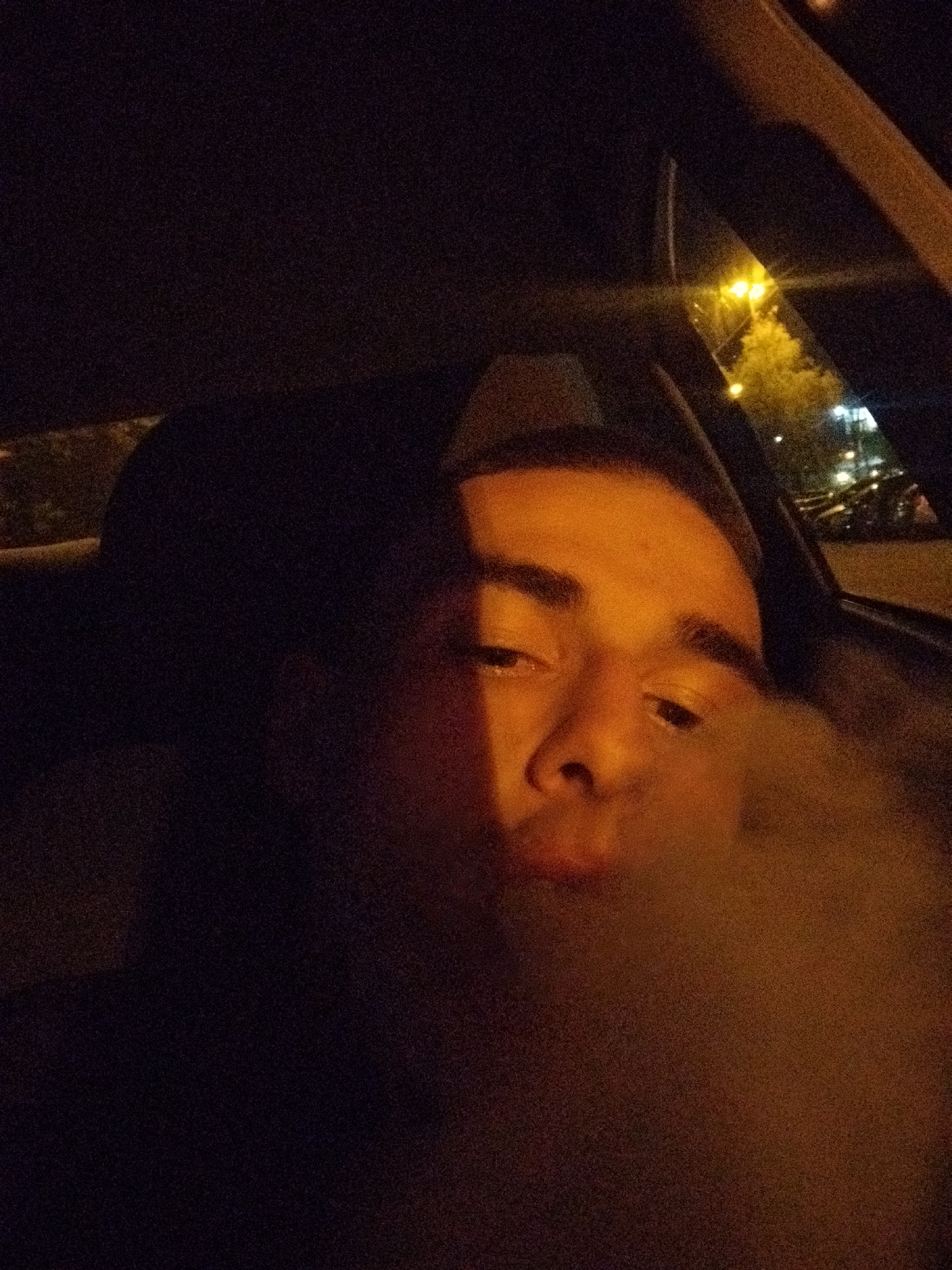 A person is sitting in the driver's seat of a car at night, blowing smoke. Streetlights are visible through the car window. It's not possible to determine the specific location in Sofia, Bulgaria without additional visual cues or information.