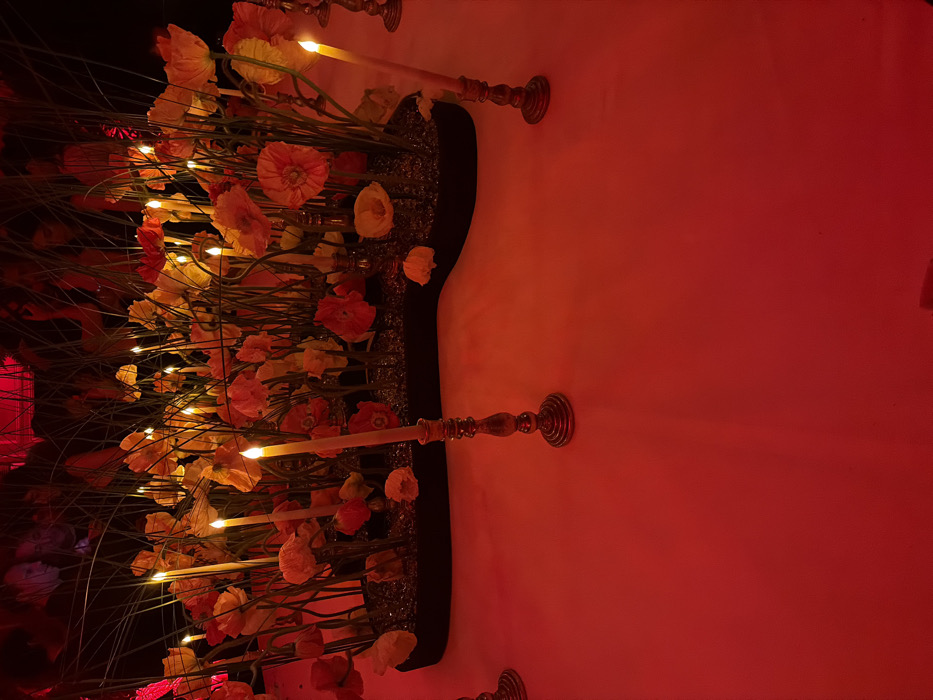 The image captures an indoor event setting, characterized by dramatic red ambient lighting. In the foreground, a smooth surface, likely a tablecloth, is bathed in deep red light. Several ornate, dark-toned metal candlesticks, possibly bronze, are visible, each holding a tall, slender, lit candle with a warm flame.
Dominating the midground is a large, irregularly shaped dark planter serving as a centerpiece. It is densely filled with a vibrant arrangement of numerous artificial or real flowers, predominantly in shades of orange and yellow, resembling poppies or similar large-petalled blooms. These flowers are interspersed with thin, tall green reeds or grasses and small, reflective granular material covering the planter's base. Several additional lit candles, similar to those in the foreground, are integrated within the floral display on thin stalks, casting a warm glow amidst the blossoms.
The background is dark and largely out of focus, revealing vague, dark silhouettes of multiple individuals, suggesting the presence of attendees at a social gathering or event. The overall environment conveys an intimate and decorative atmosphere. No specific architectural or landmark details are present to precisely identify the location within Paris.