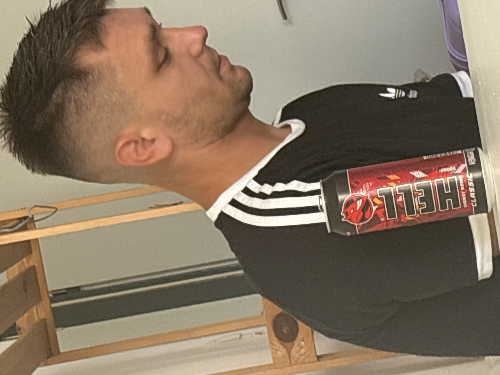 This is a vertical, close-up photograph of a man from the chest up, seen from the side. He is wearing a black t-shirt with white stripes on the sleeves, similar to an Adidas design. His eyes are closed, and his head is tilted back slightly. He has short, dark hair and stubble.
Attached to his t-shirt, on the right side of the frame, is a red can of "HELL ENERGY DRINK CLASSIC". The can features a cartoon devil logo and bold white lettering. The man appears to be indoors, with a plain wall or background behind him. A wooden shelf unit is visible in the lower portion of the image. The lighting suggests it might be daytime, but there are no clear indicators of the specific time of day or weather conditions. The man's closed eyes and relaxed posture could suggest a moment of rest or contemplation.