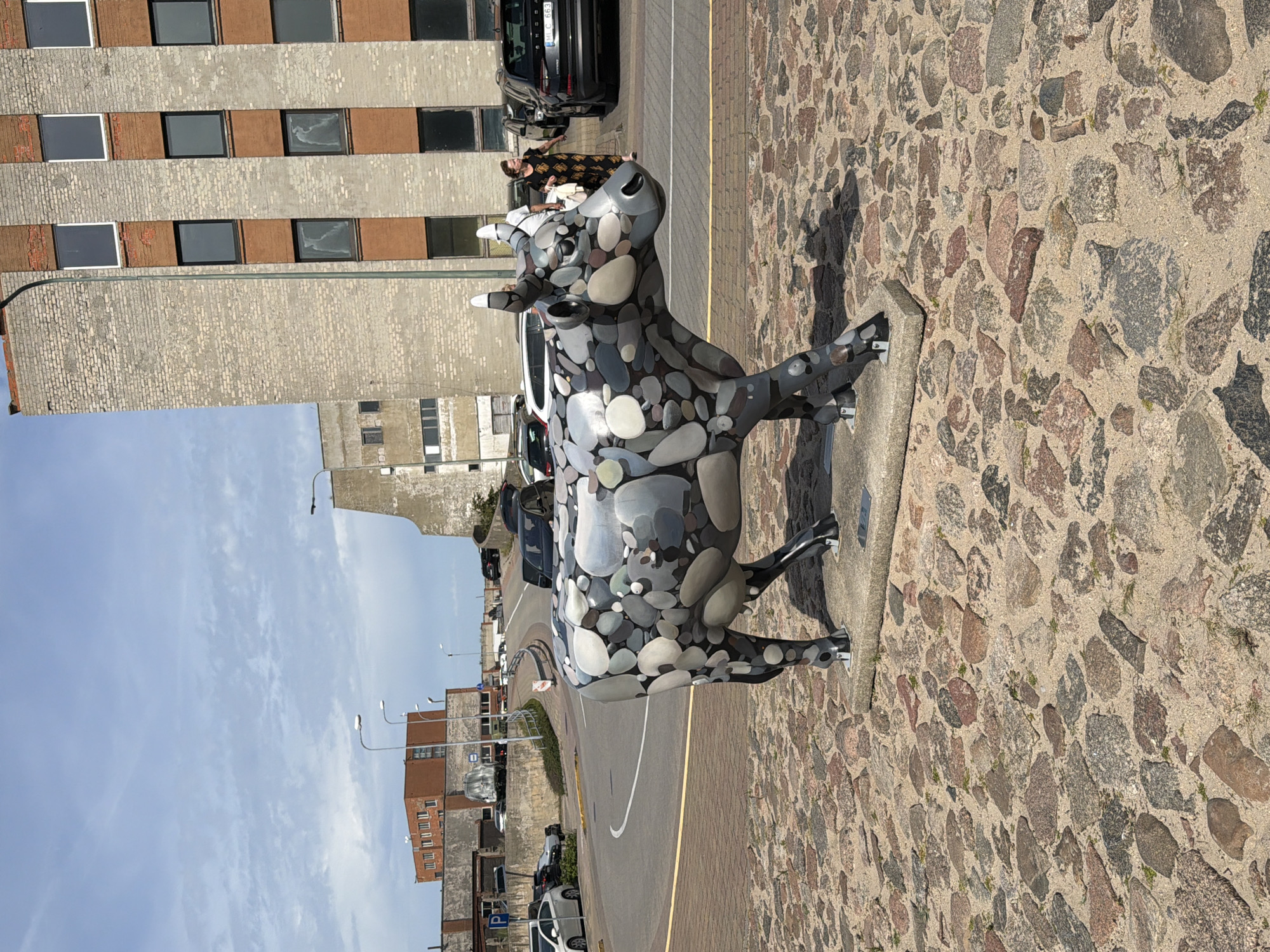 In Ventspils, Latvia, a stone mosaic sculpture of a cow stands on a cobblestone surface in front of a modern brick building. The cow sculpture is detailed, with its body made of various sized and colored stones. The building behind it has a brick facade with dark windows and some sections of reddish-brown brickwork. A street with cars and streetlights curves in the background, leading towards other buildings. A woman in a patterned dress walks away from the camera near parked cars. The sky is blue with scattered clouds.