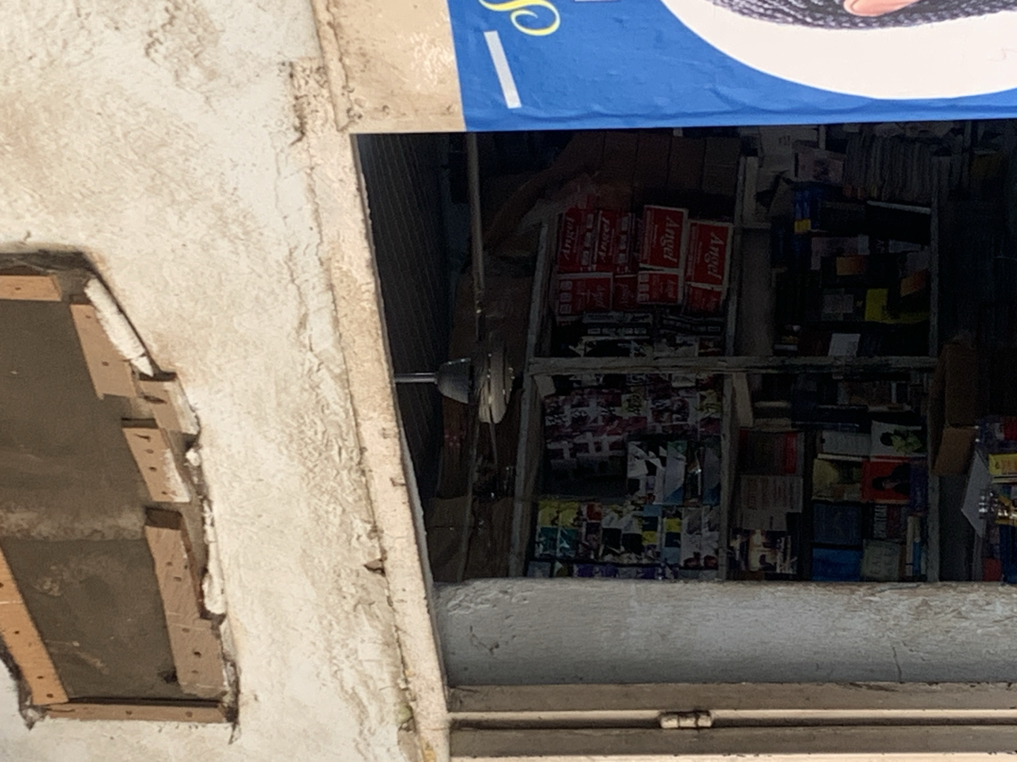 The image shows the exterior of a shop, likely a bookstore or convenience store, in an urban setting. The facade of the building is constructed of a rough, textured white material, with a wooden opening above the storefront. The opening appears to have been patched or reinforced with wooden planks. A dark-colored ceiling fan is visible hanging inside the shop.
The storefront itself is an open doorway, revealing shelves packed with various items. Stacks of what appear to be books, magazines, or other paper goods are visible on the shelves, with colorful covers and spines. Some boxes with the word "Angel" printed on them in red lettering are also stacked on the shelves. A large blue poster with a white diagonal stripe and a partial image of a person's head is affixed to the right side of the storefront, partially obscuring the view inside. The overall impression is one of a cluttered, active retail space. The lighting suggests it is daytime.