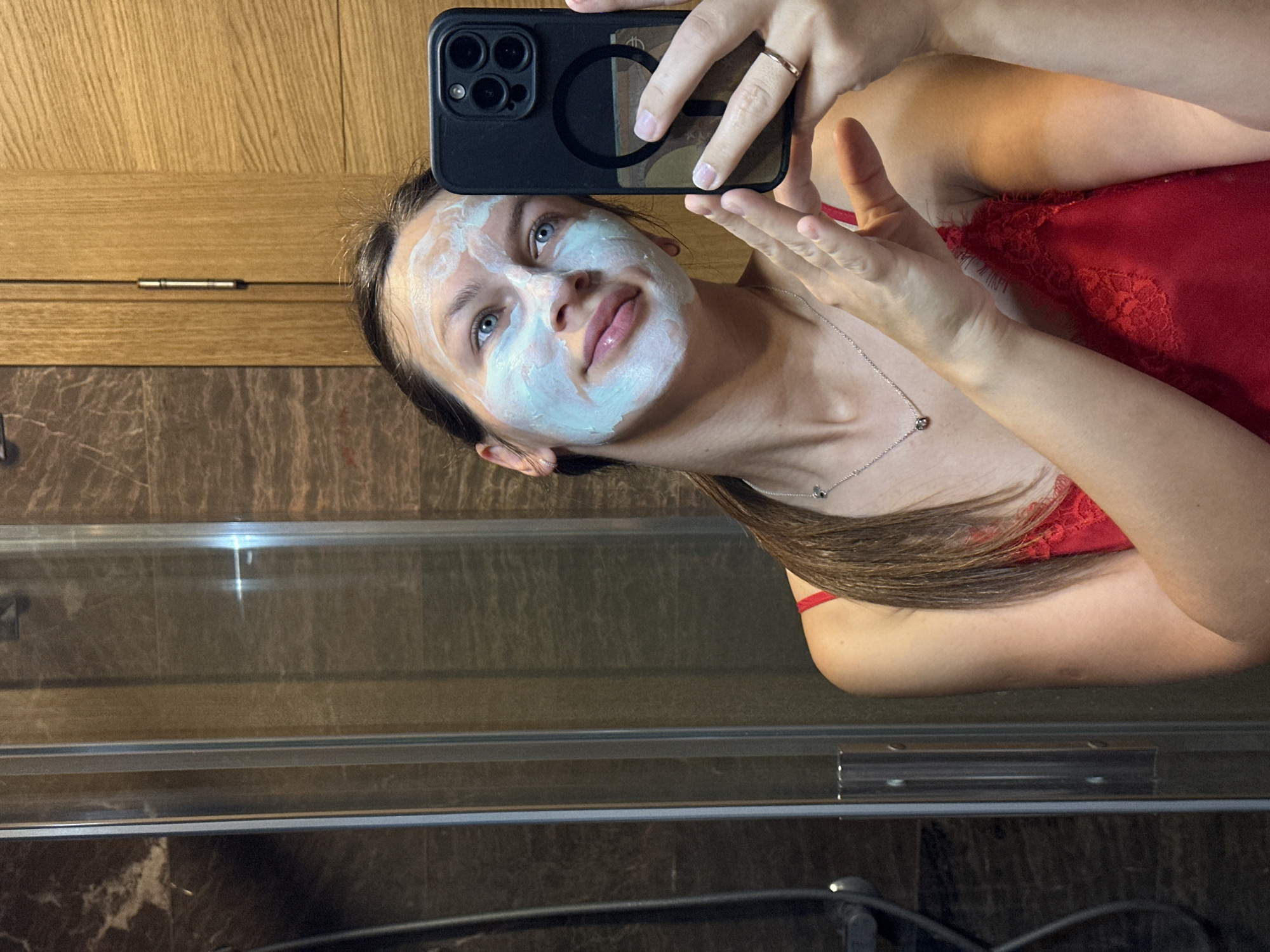 A young woman with blue eyes and brown hair, her face covered in a light blue face mask, takes a selfie in a bathroom. She is wearing a red lace camisole and a silver necklace. She holds a black smartphone in her right hand. The bathroom appears to have marble and wood paneling on the walls, and a glass shower door is visible to the left. The scene is likely in the city of Lapčići, Montenegro, based on the provided context.