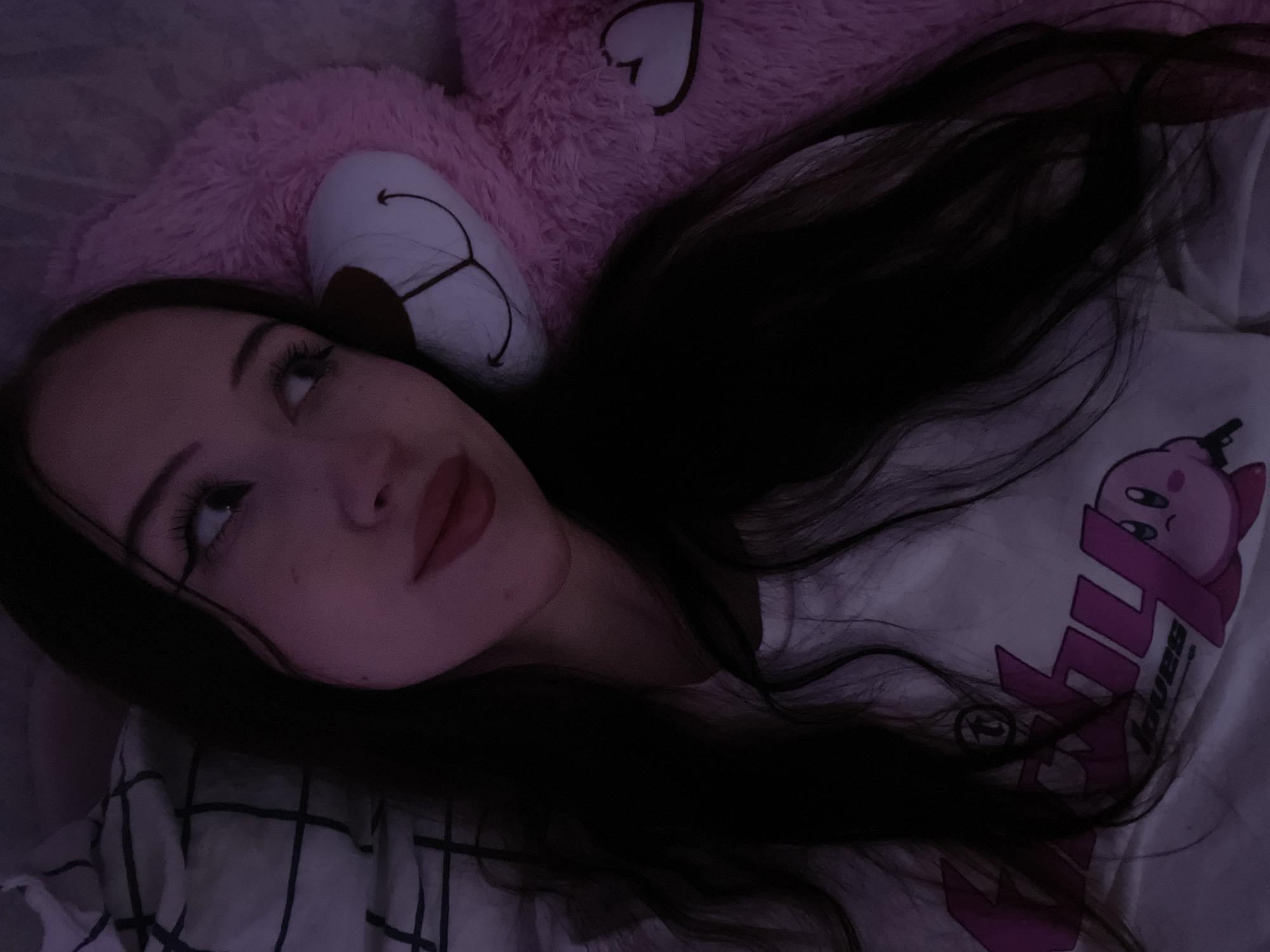The image shows a young woman lying in bed, with her head resting on a pillow and a large pink teddy bear. She is wearing a white t-shirt with a pink graphic print featuring the character Kirby. The room is dimly lit, with a purple hue cast by an unseen light source. The woman has long, dark hair and is looking away from the camera. The teddy bear is positioned behind her head and shoulder. The overall impression is one of a casual, intimate moment. This scene could be located in a bedroom within a residential apartment or house in the city of Votkinsk, Russia.
