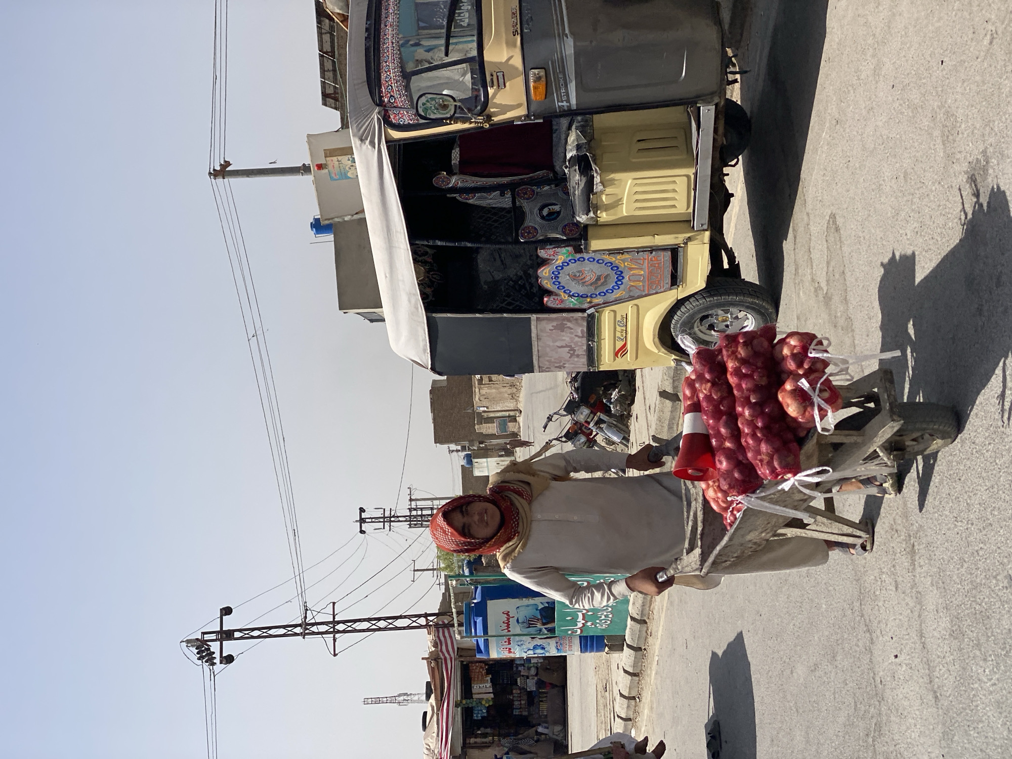 A young person wearing a red and white patterned headscarf and a light-colored tunic is pushing a wheelbarrow loaded with netted bags of red onions down a paved street in Kandahar, Afghanistan. The wheelbarrow also contains a red megaphone. To the right of the wheelbarrow is a yellow and grey auto-rickshaw, decorated with colorful patterns. In the background, there are buildings constructed of mud brick, shops with awnings, and utility poles with power lines. A blue water tank is visible near one of the shops. The sky is clear and blue, indicating a sunny day. The scene appears to be a street market or a commercial area within the city.