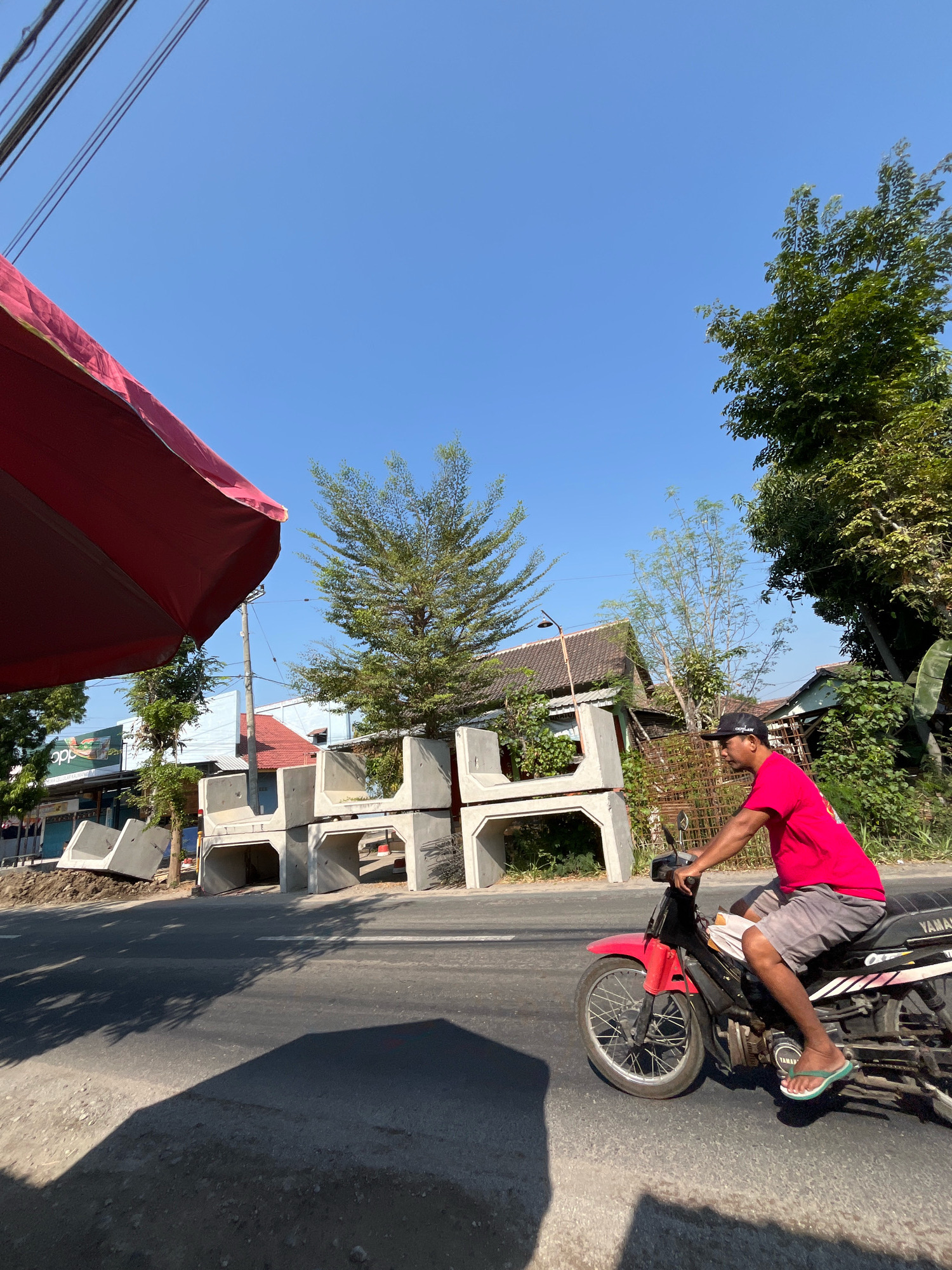 A man wearing a pink t-shirt and grey shorts rides a red Yamaha motorcycle on a paved road in Sidorejo, Indonesia. He wears a black baseball cap and green flip-flops. In the background, stacks of concrete culverts are visible on the side of the road, along with trees and buildings. A red umbrella is partially visible in the foreground, and power lines are strung across the sky. The weather appears to be sunny with a clear blue sky.