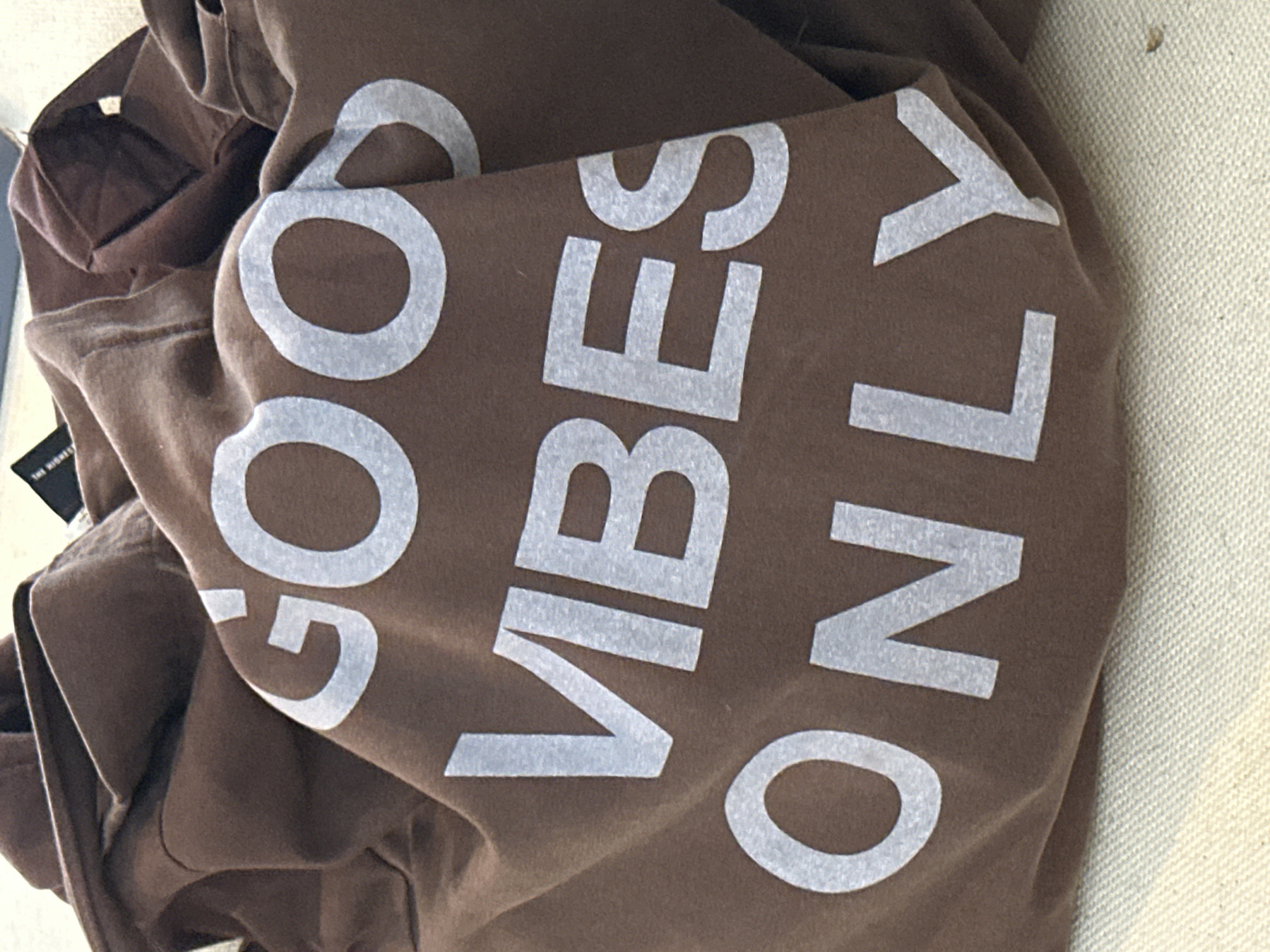 A brown t-shirt with the white text "GOOD VIBES ONLY" printed on it is presented in the image. The shirt is crumpled and lying on a textured light-colored surface. A tag is visible near the upper left corner of the shirt. The photograph appears to be taken indoors. There is no indication of the location of North Kuta, Indonesia in this image.