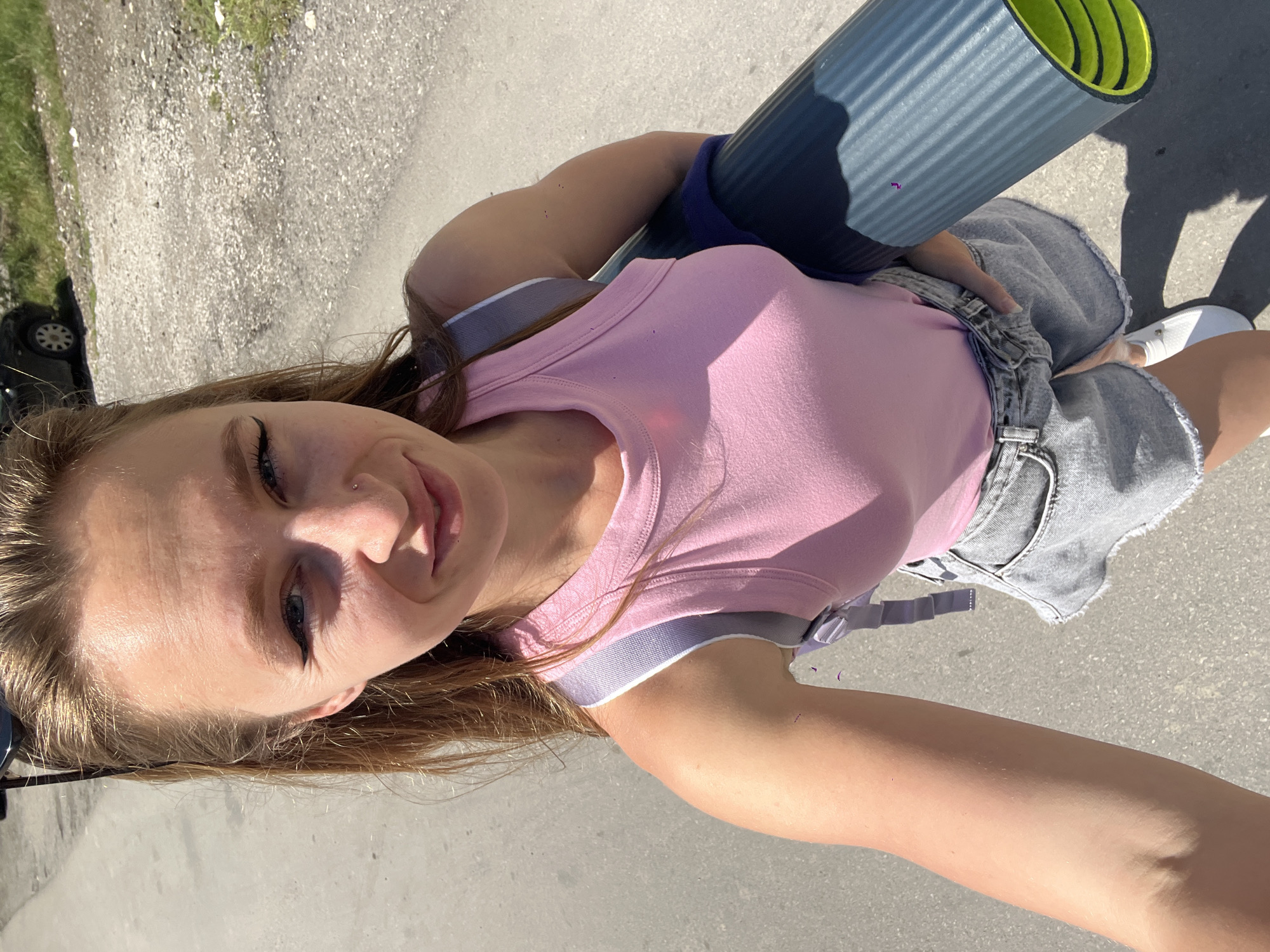 A young woman is captured in an outdoor selfie, taken in Boreti, Montenegro. She is looking upwards into the camera with a gentle smile, her long brown hair flowing around her shoulders. Her blue eyes are accentuated with eyeliner, and she has a small piercing on her nose.
She is wearing a light pink tank top over a grey and white striped sports bra. Her lower attire consists of a light-wash denim mini-skirt with distressed edges, and white sneakers on her feet. Under her left arm, she holds a rolled-up yoga mat, which is grey on the outside and bright lime green on the inside. A grey strap for the mat is visible over her shoulder.
The setting is a paved surface, likely a road or path, under bright sunlight, which casts strong, clear shadows. In the background, there's a strip of green grass and the partial view of a black car, possibly an SUV, on the left side. The bright illumination indicates it is daytime, likely midday or early afternoon, with clear weather. Her expression suggests a calm and content mood. No text is visible in the image.