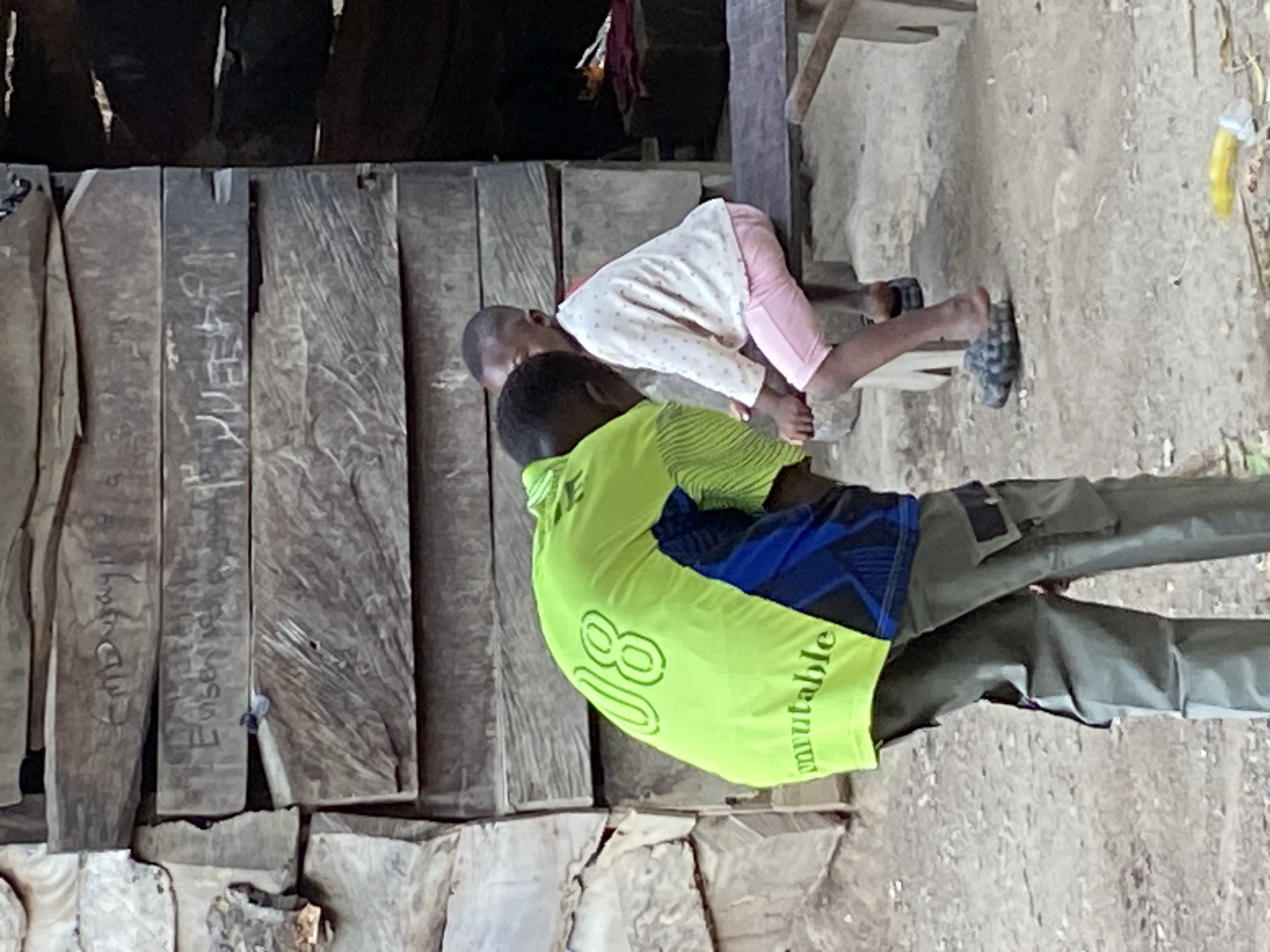 This image, taken in Mararaba, Nigeria, is rotated 90 degrees clockwise, depicting three individuals in what appears to be an outdoor or semi-outdoor setting during the daytime.
In the foreground, a person with dark skin stands with their back largely to the camera, wearing a bright neon green long-sleeved shirt. The word "Immutable" is printed vertically on the right side of the shirt's back, and the number "08" is visible on the left. They are also wearing dark, possibly cargo-style, trousers.
Behind the standing person, two younger children are present. One child, also with dark skin, is seen kneeling or leaning forward on all fours, wearing a white long-sleeved top with small pink polka dots and light pink pants. This child is wearing dark slip-on shoes. The head of a second child is partially visible between the standing person and the kneeling child.
The background consists of a rustic wall or structure made from vertical wooden planks. Several of these planks bear faded carvings or inscriptions. Visible text on the wooden planks includes "MAYI", "Emanuel", and "Esther". The ground is rough and uneven, appearing to be dirt or unpaved concrete. A yellow object, possibly a banana or a toy, lies on the ground in the mid-background. Some strips of colorful fabric (red, white) hang from above the wooden structure. The scene captures a casual moment in a local environment, with clear daytime lighting.