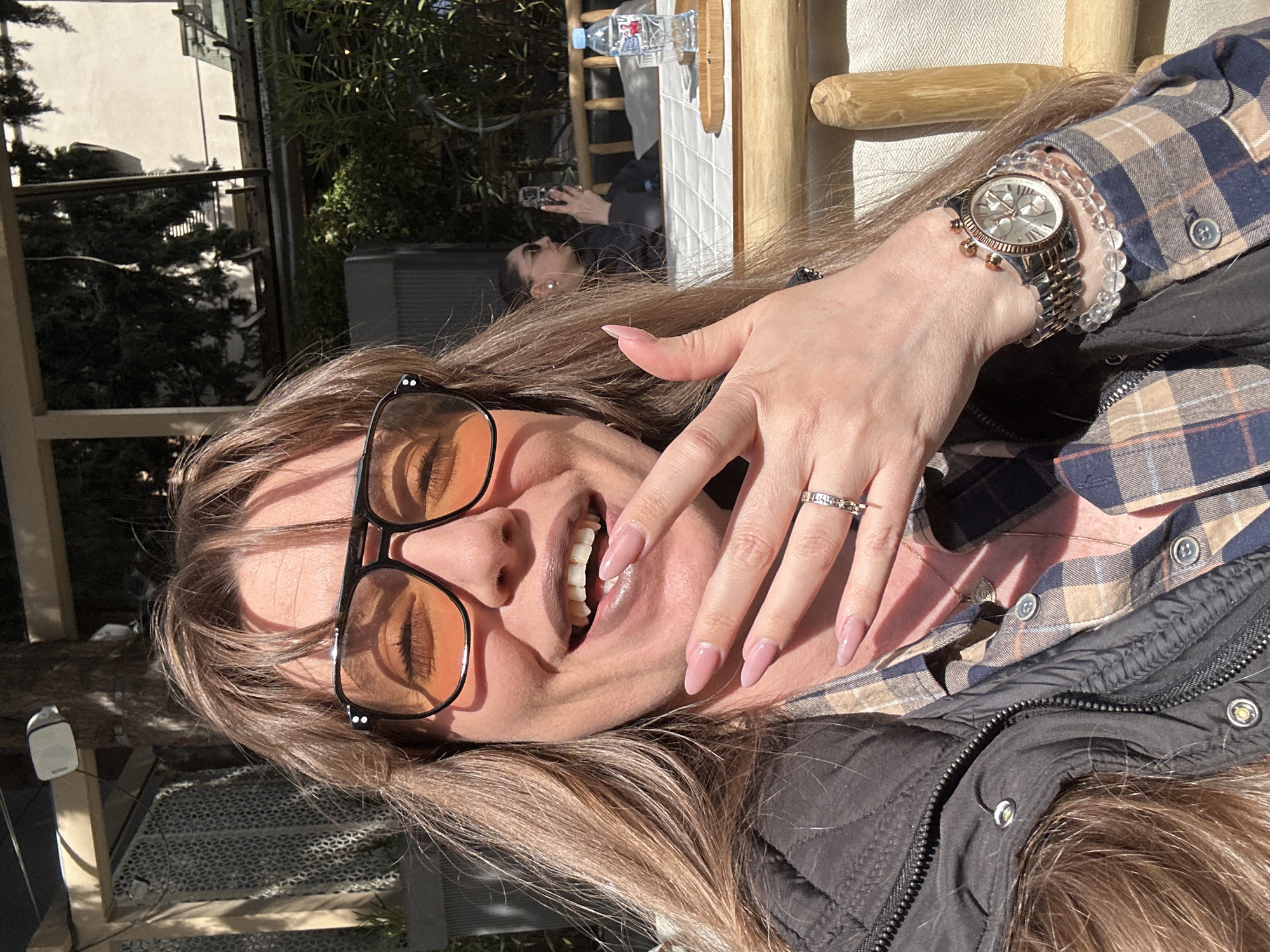 The image shows a Caucasian woman with light brown hair and glasses, smiling at the camera. She is wearing a plaid shirt under a dark jacket and has a watch on her wrist and a ring on her finger. There is a reflection of a person holding a phone in a mirror, with a water bottle above. The setting appears to be outdoors on a sunny day, possibly a patio or balcony. A building and some greenery are visible in the background, potentially indicating the city of Tbilisi, Georgia, but without additional context, it is impossible to conclusively determine the location.