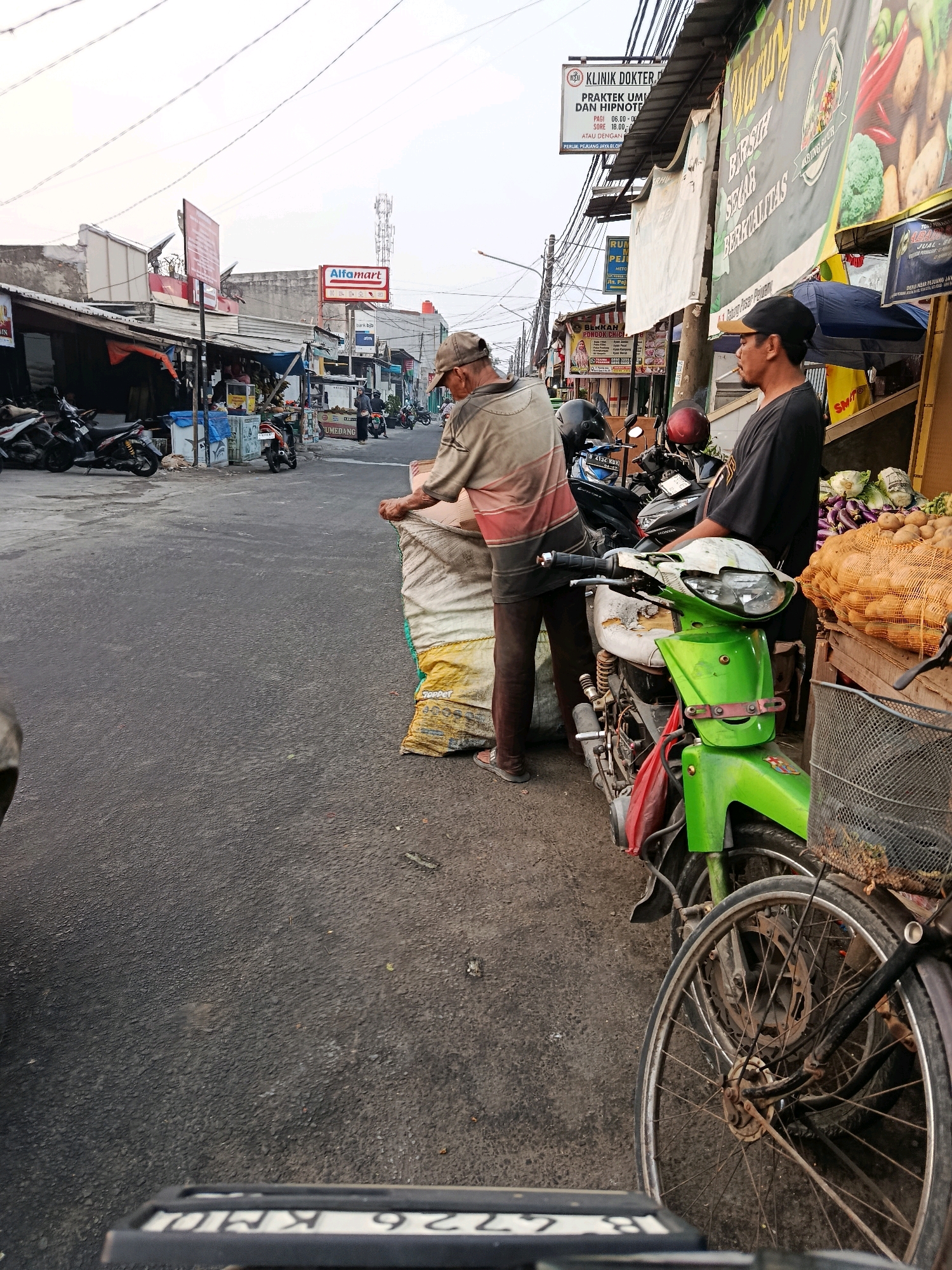 The image captures a street scene in Bekasi, Indonesia, likely on a commercial street based on the presence of shops and market stalls. In the foreground, a green motorcycle is parked, with its front wheel prominently visible. To the right of the motorcycle, a market stall displays a bounty of fresh produce, including potatoes and what appear to be cabbages or similar vegetables, arranged in sacks and baskets. Two men are present in the frame. One man, wearing a dark t-shirt and a black cap, stands next to the motorcycle. The other man, dressed in a beige shirt with red and grey stripes and brown pants, is seen from the side, appearing to adjust a large sack. In the background, several other motorcycles are parked along the street, and shop facades are visible, including an "Alfamart" convenience store sign, indicating a well-trafficked urban area. The overall atmosphere suggests a bustling marketplace environment.