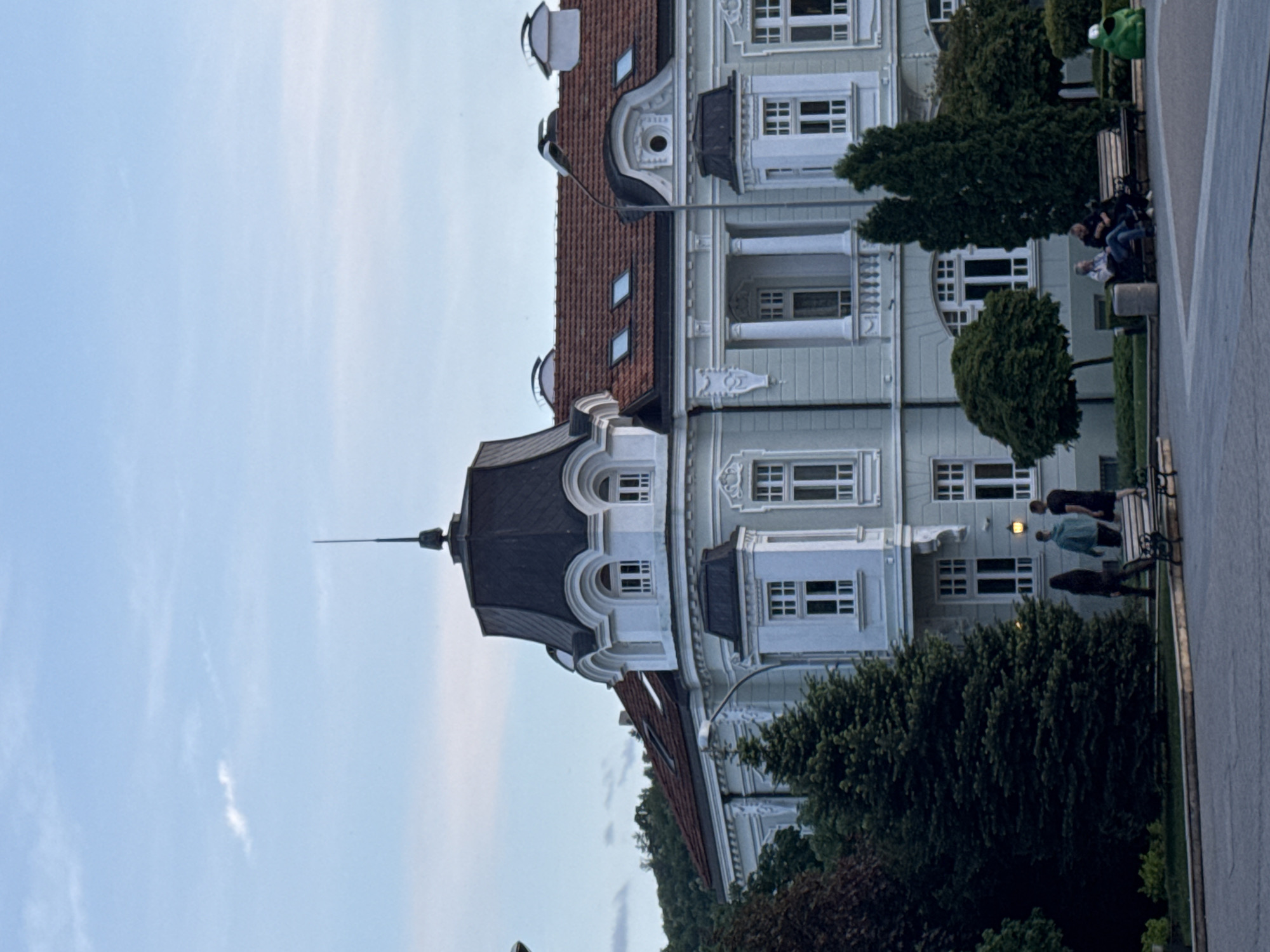The image shows a building in Unknown, Unknown. The building has a dark gray roof and light blue walls. There are white window frames and decorative elements. In front of the building, there are trees and bushes. Several people are sitting on a bench or walking along the sidewalk. The sky is light blue.