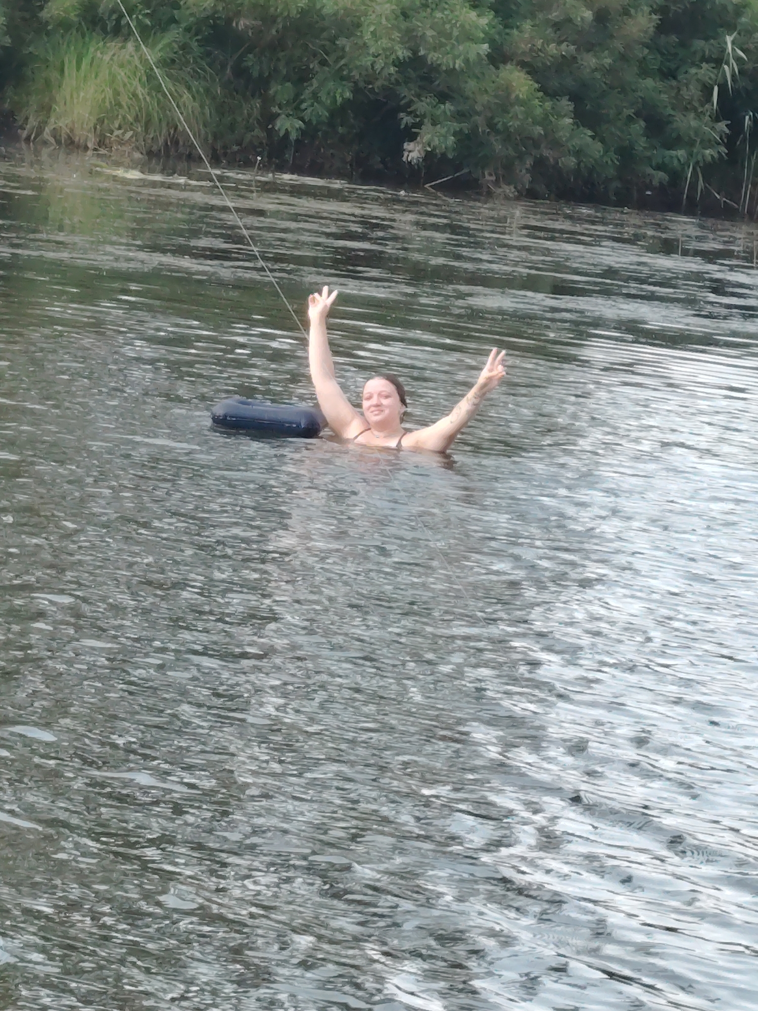 A young woman with brown hair is in the water, with a black inner tube beside her. She is wearing a dark-colored swimsuit and has her arms raised, with her index and middle fingers extended in a peace sign on both hands. Her head is turned towards the camera, and she is smiling. The water is rippling, and the background shows a dense line of green trees and vegetation. The scene appears to be a natural body of water, possibly a river or lake, in Oseshchyna, Ukraine. A thin rope extends from the upper left of the frame into the water.
