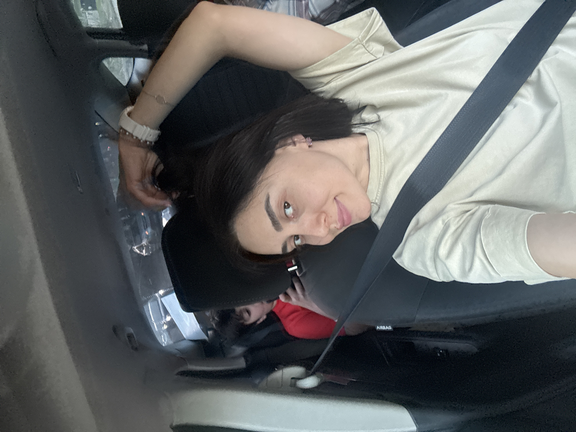 A woman is captured in a close-up shot, sitting in what appears to be the passenger seat of a car. The image is rotated 90 degrees counter-clockwise, showing her lying horizontally across the frame, but she is mentally re-oriented to be sitting upright. She has dark hair, dark eyebrows, and is looking towards the camera with a subtle, slight smile or smirk. She wears a light beige or off-white t-shirt and has a black seatbelt fastened diagonally across her chest and shoulder.
On her left wrist, she wears a white, possibly ceramic or silicone, watch and a delicate bracelet with a small, circular charm. The car's interior is visible, including a dark seat headrest and a panel marked with the word "AIRBAG." In the background, partially obscured by the seat, another individual, possibly a child, can be seen wearing a red garment. The view through the car window is blurred but suggests an urban environment, aligning with the given context of Tbilisi, Georgia. The scene appears to be taking place during the daytime.