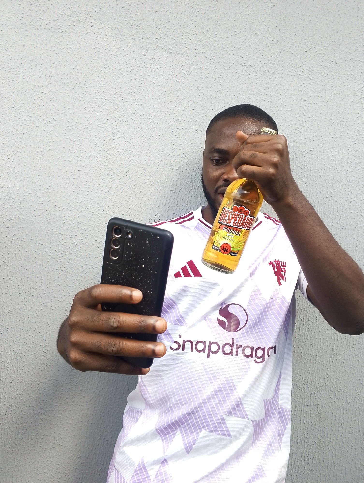 A young Black man stands in front of a textured, grey wall, holding a cell phone in his left hand and a bottle of Desperados Original Beer in his right. He is wearing a white and purple jersey with a maroon Manchester United logo and the word "snapdragon" printed on it. His right hand, adorned with a gold ring, is holding the bottle up as if to show it off. The man is looking at the cell phone, which is being held in a selfie-style position, suggesting he is taking a photo or recording a video. The overall lighting suggests it is daytime. The man has a slight smile on his face, conveying a relaxed and casual mood. The textured wall behind him indicates an outdoor or semi-outdoor setting. The context of Jos, Nigeria, suggests a warm, possibly humid, environment. The visible text on the bottle reads "DESPERADOS ORIGINAL BEER."