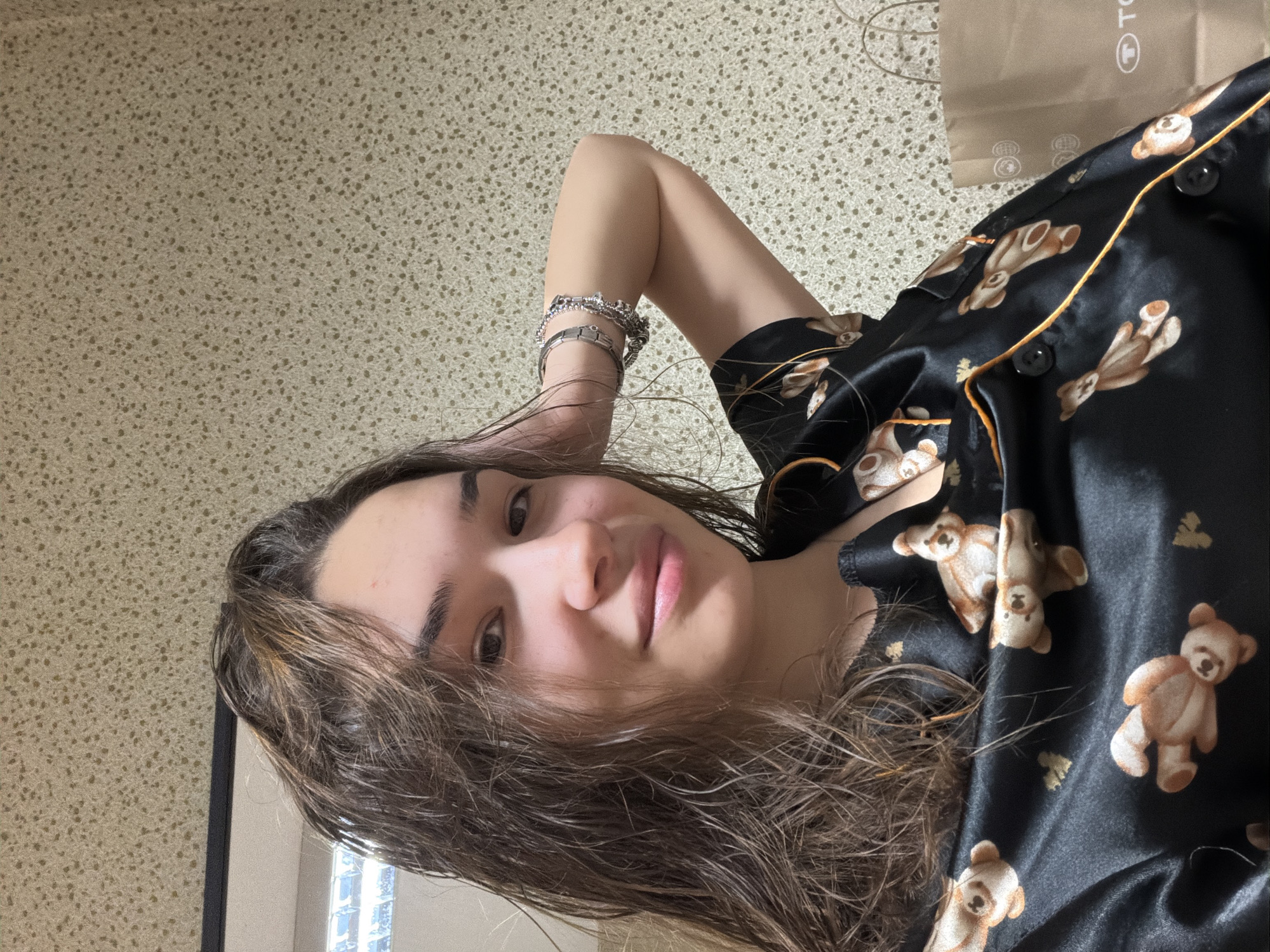 A person is seen in a close-up shot. The person is wearing a black silk pajama top with a bear print. The person has long, wet hair. The person has multiple bracelets on one arm. The location is not identifiable as Sofia, Bulgaria. The background is a beige wall. A brown bag is partially visible in the upper right corner.
