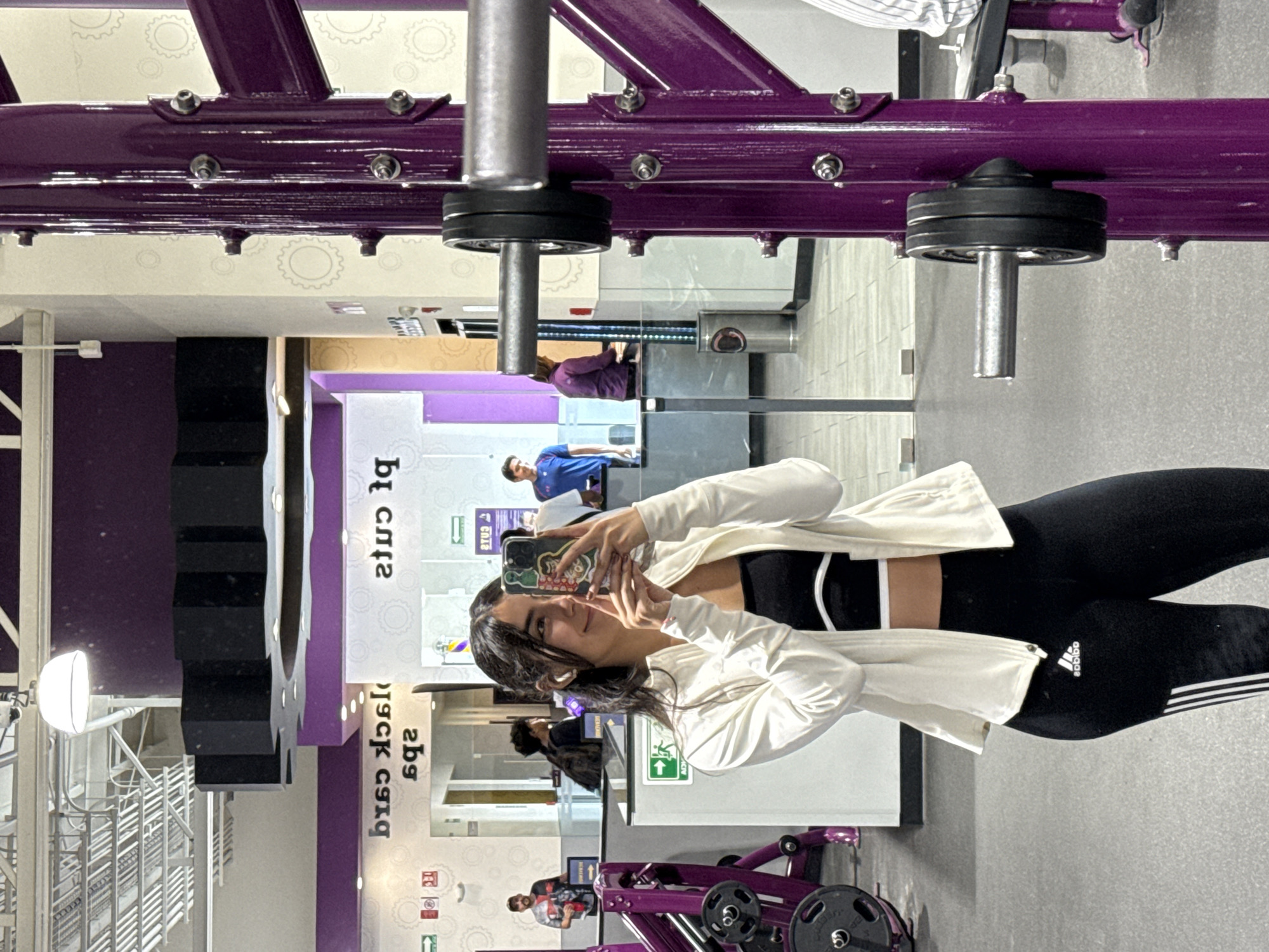 A young woman is taking a selfie in a Planet Fitness gym in Puebla, Mexico. She is wearing black Adidas leggings, a black sports bra with white trim, and a white jacket. She is holding her phone in her left hand, facing a mirror. The gym features its signature purple equipment. Other gym patrons are visible in the background.