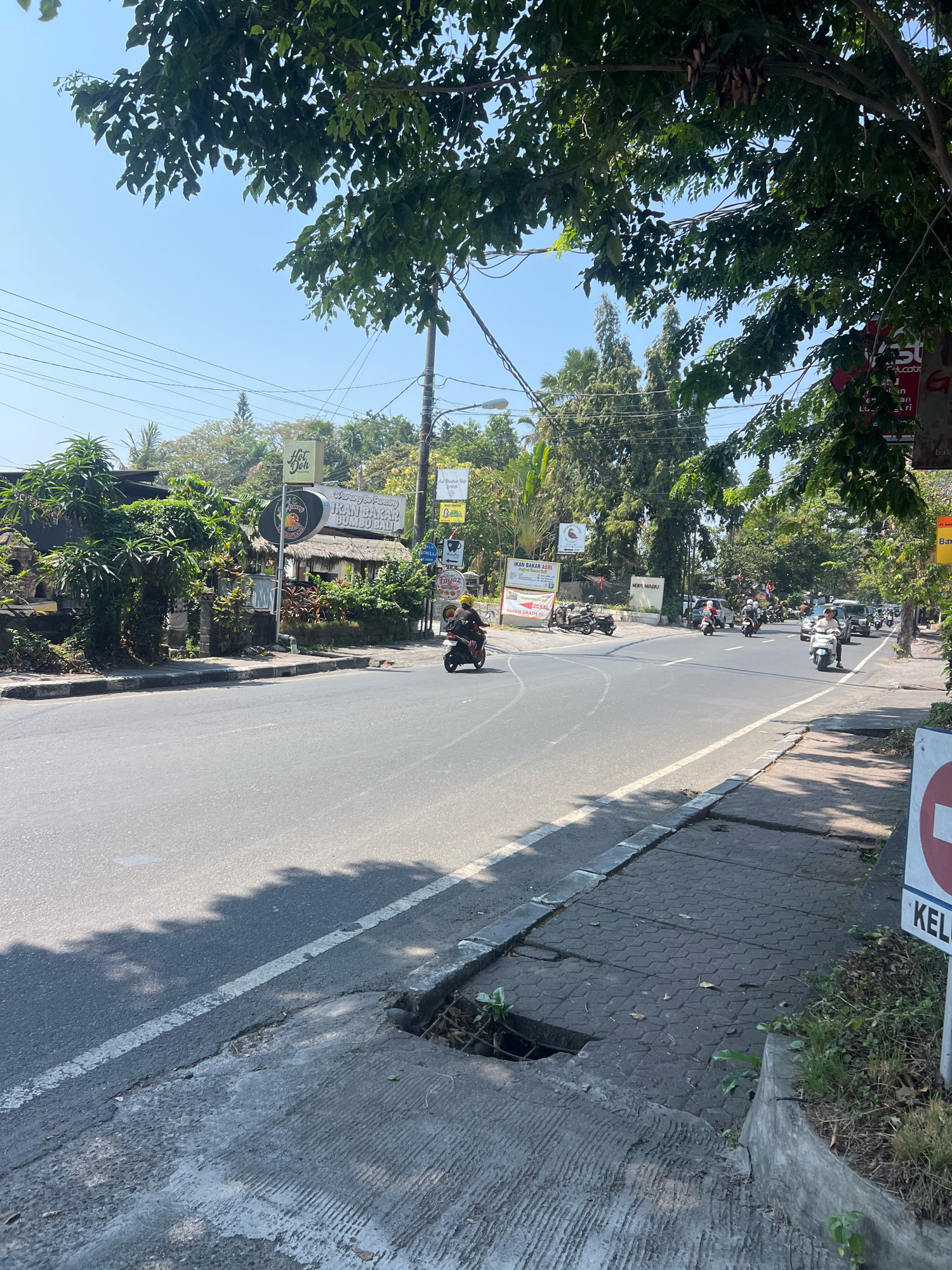 The image depicts a street scene in Denpasar, Indonesia, likely a busy area with businesses lining the road. The day is sunny with a clear blue sky. A two-lane asphalt road stretches into the distance, with a pedestrian sidewalk made of interlocking pavers on the right. There is a drainage grate on the sidewalk. Several motorbikes and a few cars are visible on the road, indicating traffic. On the left side of the road, there are trees and foliage, as well as some low-rise buildings with thatched roofs. Several signs are posted, some with Indonesian text, suggesting commercial establishments like "Warung Makan" (eatery) and "Ikan Bakar Bumbu Bali" (Balinese grilled fish). On the right side of the road, there is a sign with a red circle and a diagonal bar, a common no-entry symbol, and the word "KELUAR" (exit) below it. Overhead, power lines crisscross the sky. The overall impression is a vibrant and active urban environment in Bali.