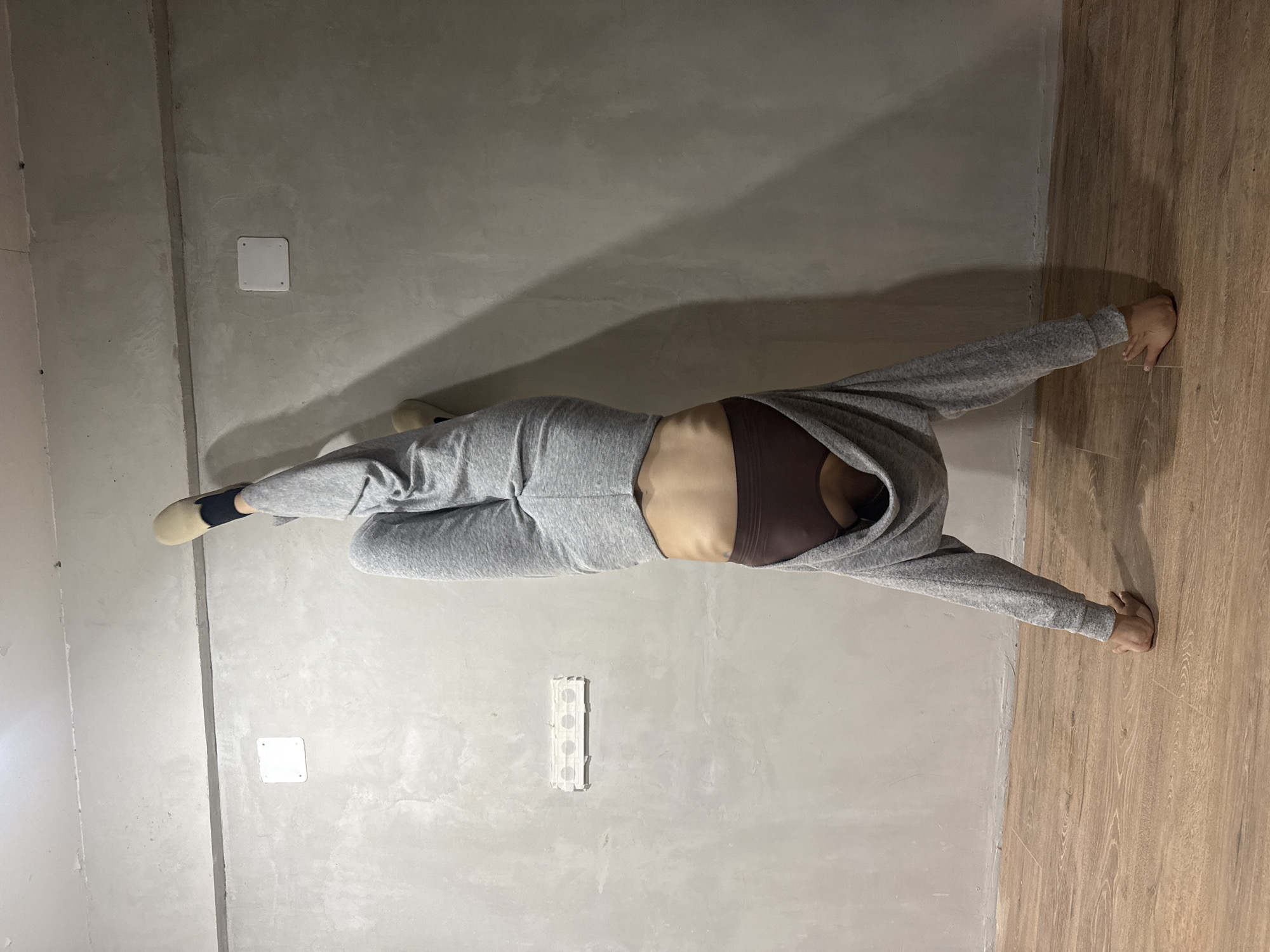 The image shows a person in a gray sweatsuit and brown sports bra lying on the floor of a room. The room has two different types of flooring. One section of the floor appears to be wood or laminate, while the other is a smooth, gray surface. The person is positioned with their legs bent to the left and their arms extended in opposite directions. The person's right hand is placed flat against the wooden/laminate floor, and the rest of their body is located on the gray floor. The person is in a position, which might be some type of yoga exercise. There are two white square covers on the gray wall. There is a long white rectangular object on the gray wall. The image does not provide any information to identify the location as Votkinsk, Russia.
