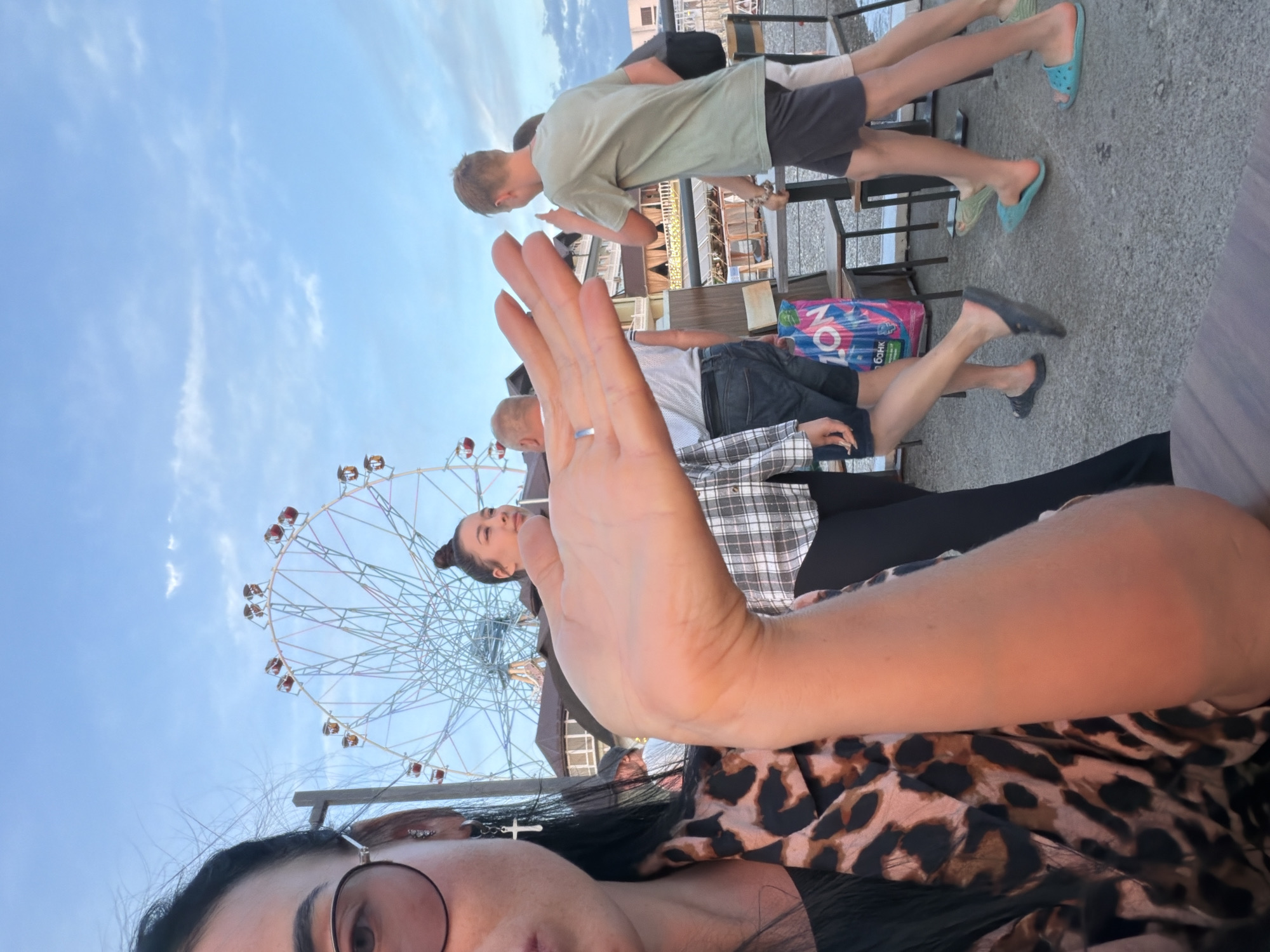 An outdoor scene at dusk in Sochi, Russia, features a prominent Ferris wheel in the background against a pale blue sky with scattered clouds. In the foreground, a woman with dark hair and glasses is partially visible on the left, extending her right hand towards the camera. She is wearing a black garment and a leopard-print jacket.
Several other individuals are in the midground. To the right of the woman with the extended hand, another woman in a plaid shirt is looking away from the camera. A young man wearing a green t-shirt and dark shorts walks past, gesturing with his right hand. Further to the right, another person is partially visible.
The environment appears to be an open area, possibly a promenade or a park, with tables and chairs set up in the midground. A pink shopping bag with Cyrillic text is on the ground near the tables. The ground is paved with a rough, grey surface. The overall lighting suggests it is late afternoon or early evening.