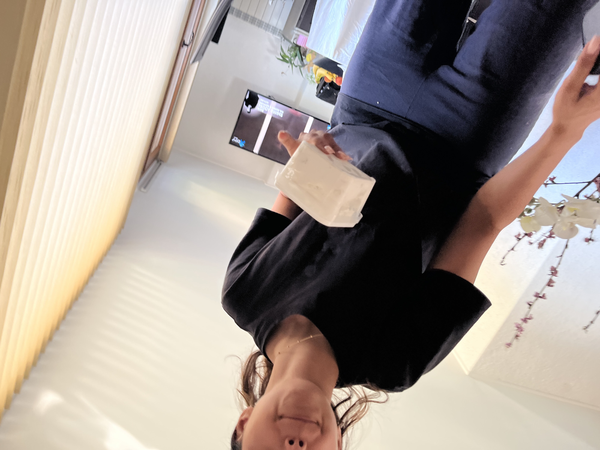 An indoor scene in Zlatograd, Bulgaria, captures a young adult female from a low-angle perspective. She is positioned slightly to the right of the frame's center, looking towards the viewer with a subtle smile, her face partially obscured. She has dark hair, a delicate gold-colored beaded necklace, and a nose piercing on her left nostril. She is wearing a black short-sleeved t-shirt and dark blue or black pants. Her right hand, visible in the lower center, holds a white, rectangular block, possibly a food item like butter or cream cheese, and her thumb is extended upwards in a "thumbs up" gesture.
The background reveals a domestic setting. To the right of the individual, a large flat-screen television is visible, displaying what appears to be a broadcast with a "City" logo and vertical white bars on the screen. A green potted plant with elongated leaves stands near the television. Further to the right, a white-sheeted surface, possibly a bed or table, is present. In the bottom right foreground, a floral arrangement featuring white orchids and thin branches with small reddish-purple buds is visible. The upper portion of the image shows light-colored horizontal window blinds or shades, through which sunlight casts distinct striped shadows onto a white or off-white wall. A slender, metallic rod is visible below the blinds. The overall lighting suggests a bright interior space with natural light.