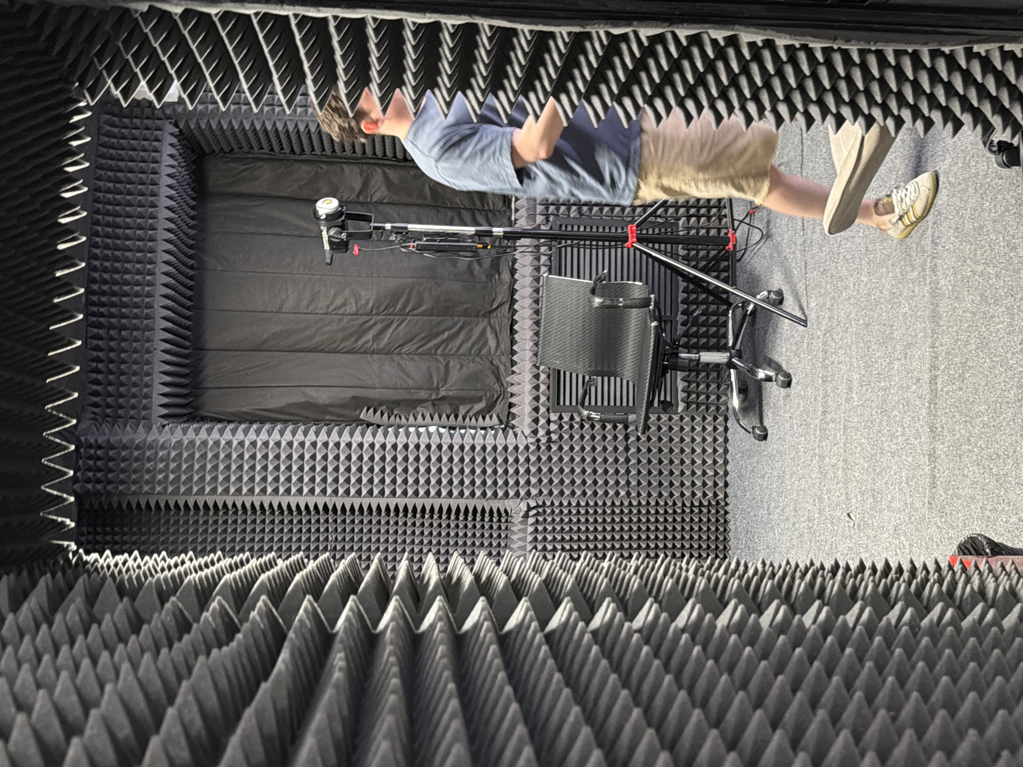 This image depicts the interior of a soundproof room, likely a recording studio or podcast booth. The walls, ceiling, and floor are covered with dark grey acoustic foam panels featuring a pyramidal texture. In the center of the room, a black office chair with wheels is positioned. To the right of the chair, a tripod with a light fixture and cables attached stands. A person, partially visible from the waist down, is walking through the room from right to left. They are wearing a blue t-shirt, khaki shorts, and light-colored athletic shoes with yellow stripes. The overall environment is dark and enclosed, designed to minimize external noise and reverberation. There is no identifiable information within the image that would indicate a specific location in Moscow, Russia.