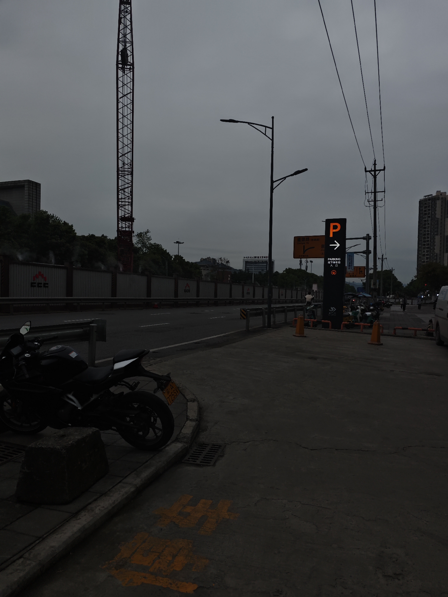 The image depicts a street scene in Chongqing High-tech Zone, China, on a cloudy day. A black motorcycle is parked on the sidewalk next to a low concrete barrier. Beyond the sidewalk is a street with a metal barrier along its edge. Behind the barrier is a construction site with a tall crane in the background. A parking sign with a glowing orange "P" and an arrow pointing right is visible on the sidewalk. Streetlights, power lines, a building, and a parking lot are also visible in the background. People, motorcycles and a vehicle are parked along the street. Yellow markings are visible on the sidewalk in the foreground.