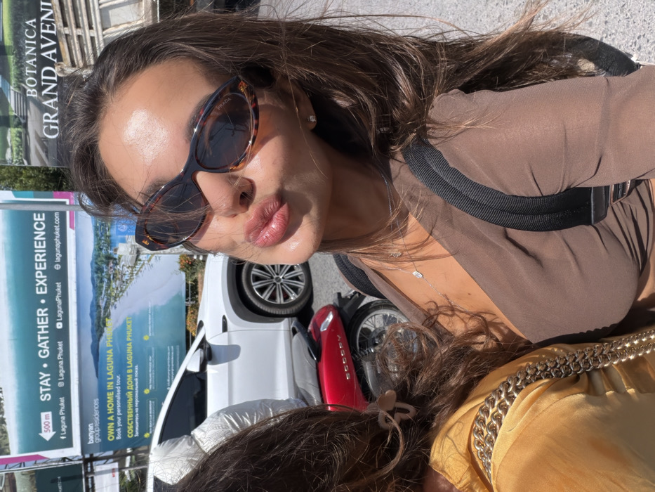 A woman wears sunglasses and poses for a selfie near a billboard advertising Laguna Phuket residences. A white car and a red scooter are parked nearby. She is wearing a brown top with black accents and a delicate necklace. Part of another person wearing a gold top with a large chain is visible at the bottom of the image. The billboard reads "Stay, Gather, Experience" and features the Botanica Grand Avenue logo. The woman's sunglasses appear to be a Prada brand.