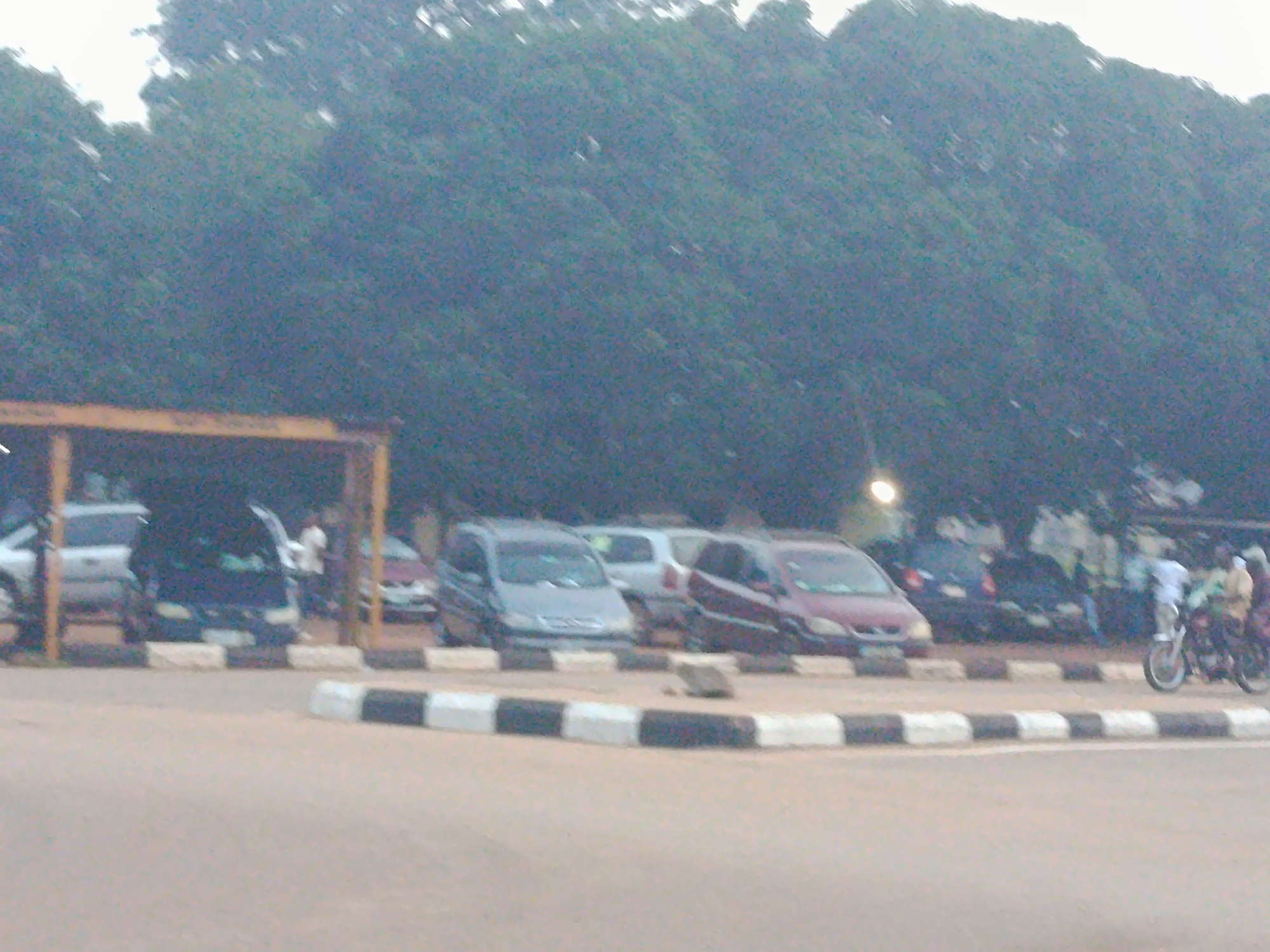 A low-angle, slightly blurred outdoor photograph captures a street scene with several vehicles and a few individuals in what appears to be Maigizo, Nigeria. The foreground features a paved road bordered by a concrete curb with alternating black and white painted blocks. Behind the curb, a row of parked cars is visible.
On the left, a white SUV is partially obscured by a yellow overhead structure. To its right, a dark-colored SUV is parked, and a person in a white shirt is visible near its open door, appearing to interact with it. Further to the right, a maroon car and a silver car are parked side-by-side.
In the background, a dense line of trees forms a backdrop. To the far right of the image, two motorcycles are parked, and two individuals are standing nearby, one on each motorcycle. The lighting suggests it might be late afternoon or early evening, with a faint light source visible in the upper left portion of the background. The overall scene depicts a typical street or parking area with everyday activities.