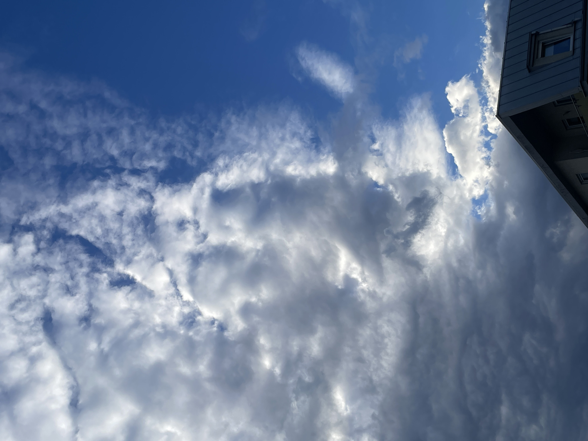 The image shows a view looking up at a sky with dynamic clouds and the roofline of a building. The sky is a vibrant blue with a mix of dark, heavy clouds and lighter, wispy ones. The clouds are textured and varied, with some areas appearing bright from sunlight filtering through, while others are shaded and dense. The building's roof is made of dark, possibly metal, material and has a distinct angular shape. A small window is visible on the slanted roof. The overall impression is of a dramatic sky with an urban element.
