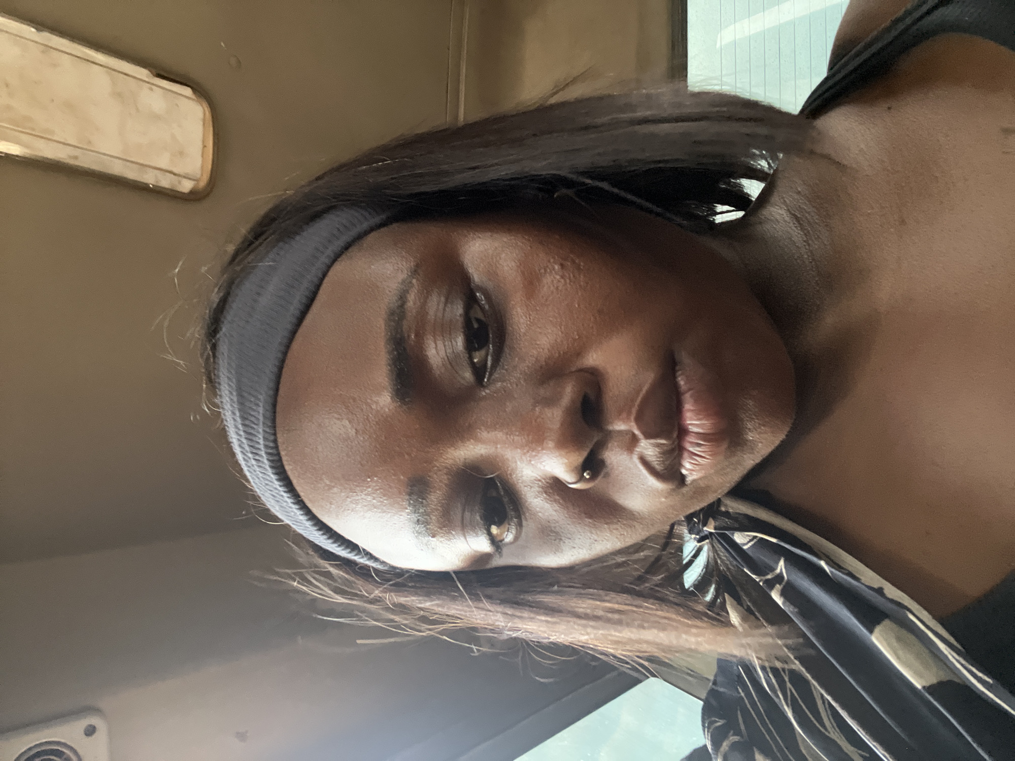 A woman is captured in a close-up, selfie-style shot, looking down. She has dark skin, dark hair, and is wearing a dark grey headband. A small stud is visible in her nostril. She is wearing a black garment with a white and black patterned fabric draped over her shoulder. The background appears to be the interior of a vehicle, with a light-colored panel and a light fixture visible on the ceiling, and a window with vertical lines on the right side. The image quality is moderate, with some visible skin texture and slight blurring, likely due to the angle and movement of the selfie. The lighting is natural and appears to be coming from the side, highlighting the contours of her face. The scene suggests a casual, on-the-go moment.