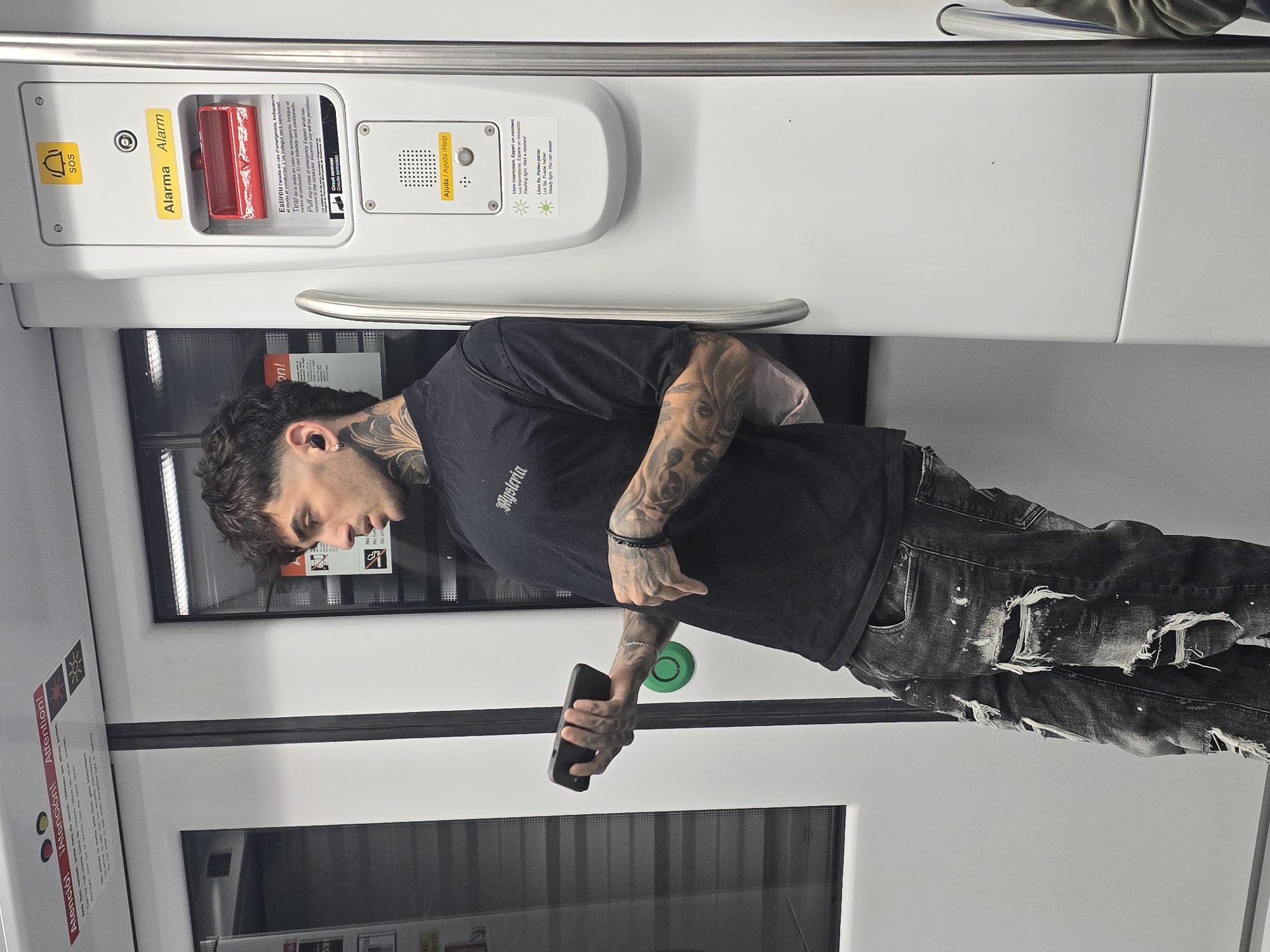 A photograph captures a person with tattoos and a mullet haircut inside what appears to be a Barcelona Metro train car. He is wearing a black t-shirt with the word "Mysteria" on the front, and torn jeans. He is holding a cell phone in his left hand. He is leaning against a hand rail.
The train car interior features white walls with emergency pull rings labeled in multiple languages ("Atenció! Atención! Attention!"). An intercom system labeled "Ajuda / Ayuda/Help" is visible near the top of the photograph. Safety information and instructions are presented in Catalan, Spanish, and English. The image provides a sense of confined space, typical of public transportation.