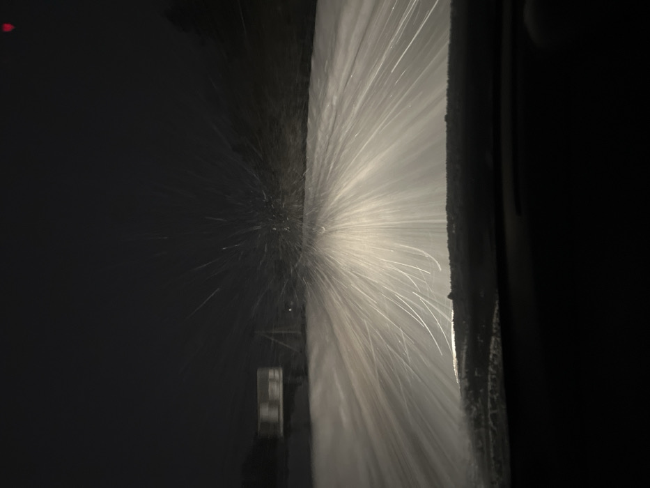 The image presents a nighttime scene in Davydkovo, Russia, captured from inside a vehicle looking through its windshield. Heavy snowfall is visible, illuminated by the vehicle's headlights, appearing as numerous bright white streaks radiating upwards and outwards from the lower-center of the frame. The road surface ahead is uniformly covered in snow. The vehicle's dark dashboard occupies the lower portion of the image, bordering a snow-dusted lower edge of the windshield. In the blurred left background, a light-colored, rectangular object resembling a roadside sign or billboard is partially visible, elevated on posts. The right background features indistinct dark shapes suggesting trees or shrubbery. A faint red light source is barely discernible in the upper right quadrant. No individuals are present within the frame.