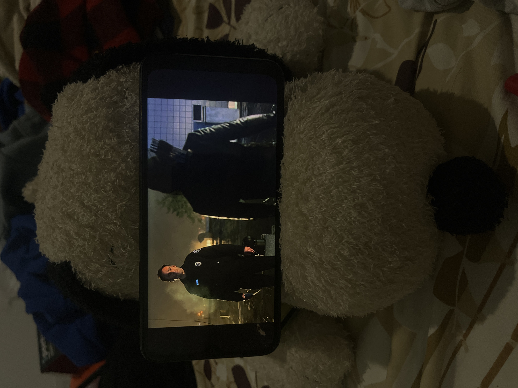 A close-up, eye-level shot shows a smartphone displaying a scene from a television show. The phone is positioned on a fluffy, light-colored stuffed animal, which rests on a bed with a patterned comforter. The screen of the phone depicts two figures. On the left, a man in a police uniform stands looking towards the camera, with a serious expression. He is holding a dark object in his right hand. Behind him, smoke or mist rises, and distant lights are visible. On the right side of the screen, another figure, silhouetted in a dark hooded costume with a quiver of arrows on their back, is seen from the back, facing away from the viewer and towards a building. The lighting on the screen suggests an outdoor, nighttime setting. The stuffed animal and the bed indicate a domestic environment. The image does not provide any information about the location in the city of Santa Ana, Venezuela.