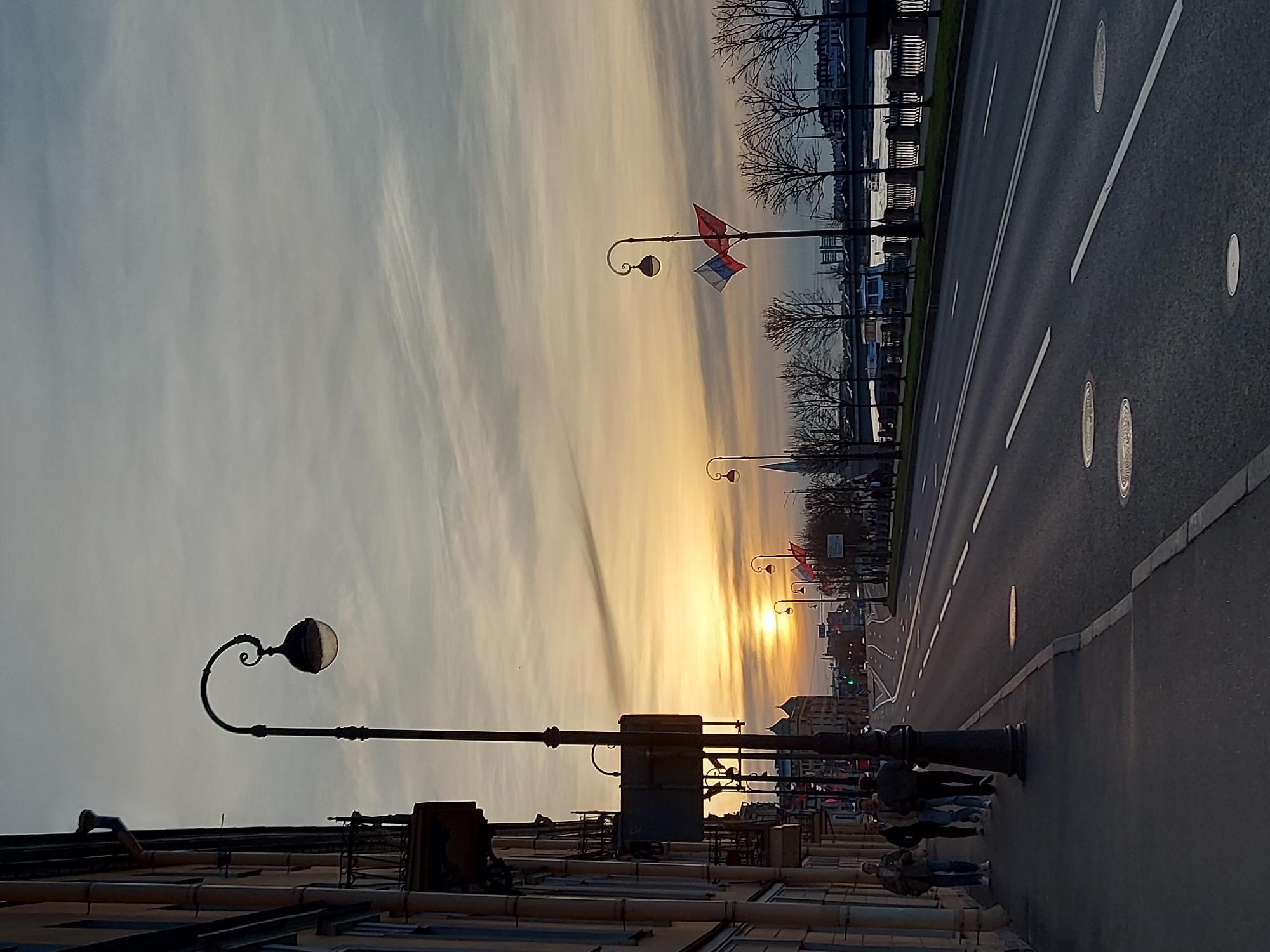 The image depicts a street scene in Saint Petersburg, Russia, during a sunset. The sky is filled with clouds and gradient colors from orange to grey. A street runs alongside a river with trees and a metal fence. There are ornate lampposts along the street. Russian flags hang from some of the lampposts. People are walking on the sidewalk. Buildings line the left side of the image, including rooftops and windows. The street has lane markings and manhole covers.