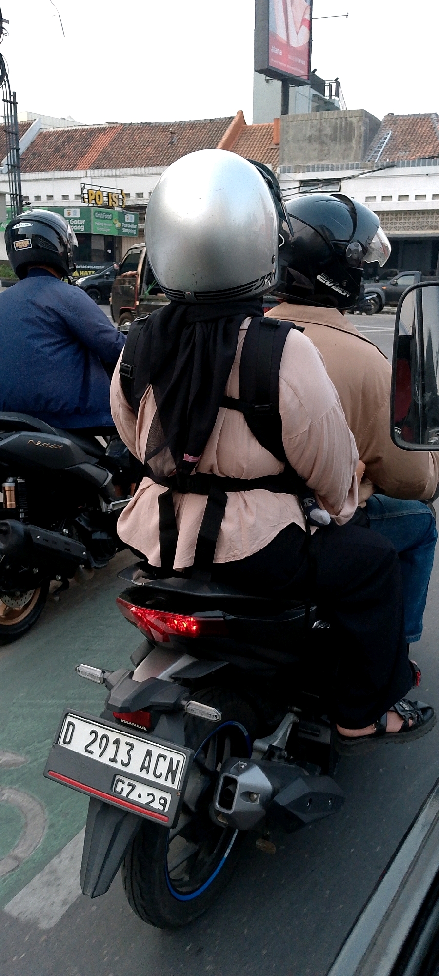 A low-angle, medium shot captures three people riding on two motorcycles in what appears to be an urban setting. The motorcycle in the foreground has a license plate reading "D 2913 ACN G7.29". The driver is wearing a dark blue jacket and a black helmet. The passenger behind the driver wears a light pink blouse, a black hijab, and black pants, and a black backpack. The third person, the driver of the motorcycle behind, wears a tan jacket and a black helmet. The background shows buildings with tiled roofs and a billboard. The lighting suggests it is either early morning or late afternoon. The presence of a vehicle with a license plate starting with "D" indicates that the location is likely in West Java, Indonesia, which includes Bandung City.