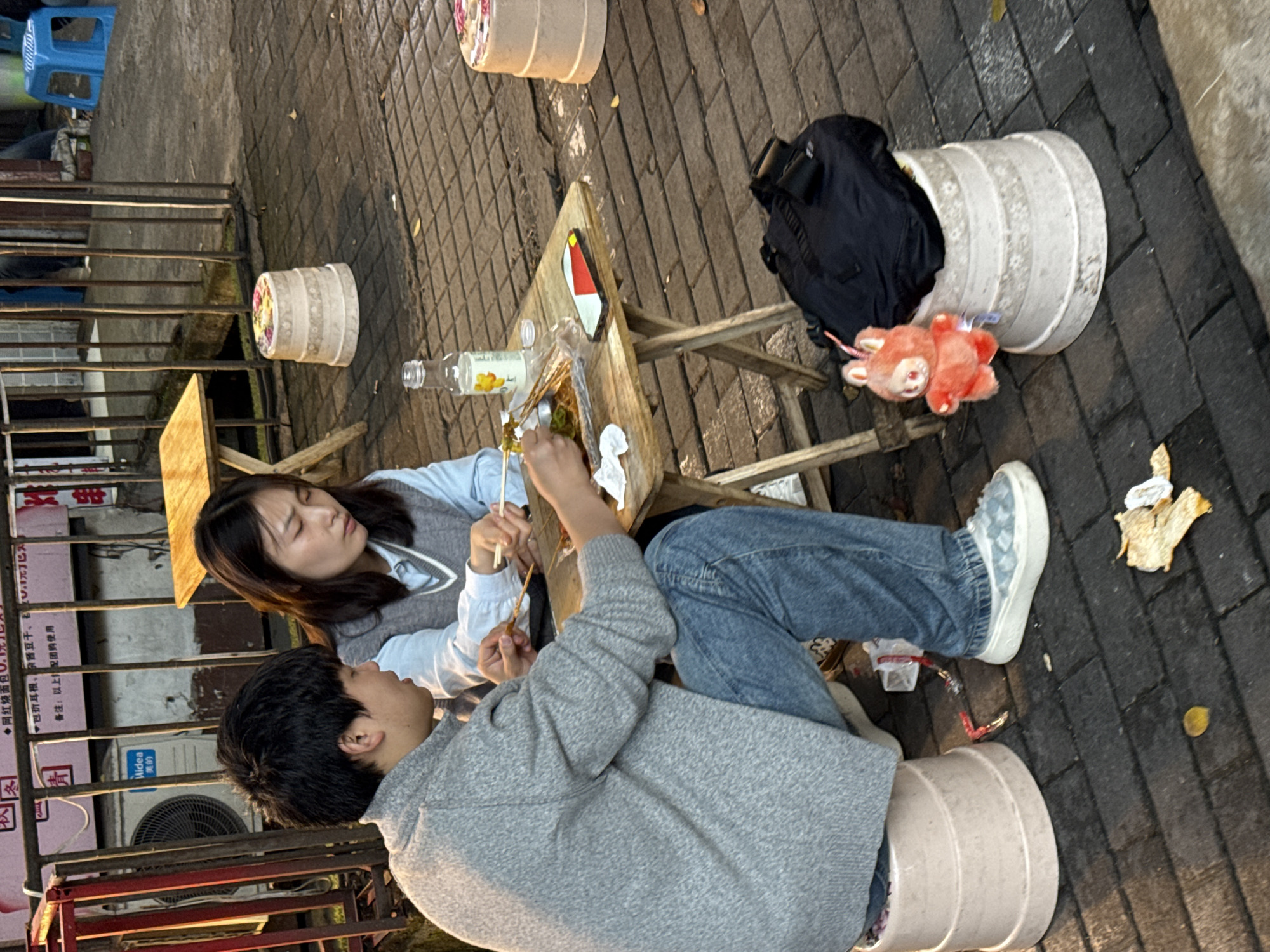 Here is a detailed description of the image:
**General Scene:**
The image captures an outdoor scene of two individuals, a man and a woman, sitting at a small, wooden table. They are eating food using chopsticks. The setting appears to be a casual, possibly street-side dining area.
**People:**
* **Woman:** The woman has dark hair. She is wearing a light blue, collared shirt, covered by a gray vest.
* **Man:** The man has dark hair and is wearing a gray sweater.
**Objects:**
* **Table:** A small, wooden folding table is positioned between the two people.
* **Food:** There are visible food items on the table, which they are eating with chopsticks.
* **Drinks:** A bottle of a light-colored liquid sits on the table.
* **Seats:** The individuals appear to be sitting on makeshift seats, which are large, white, cylindrical containers.
* **Personal Items:** There are personal items like a black bag and a plush toy located on one of the seats.
**Actions and Interactions:**
* The two individuals are actively eating food using chopsticks.
* They appear to be engaged in a meal together.
**Environment:**
* The scene is set outdoors with gray brick street.
* There are various cylindrical containers around, some appearing to be used as makeshift seating.
* An air conditioner unit is visible behind a red metal railing and a printed menu.
**Location Clues:**
Based on the Chinese characters on the menu, the presence of specific local snack foods, and other environmental details, it can be inferred that the location is most likely in the city of Yuzhong District, China.