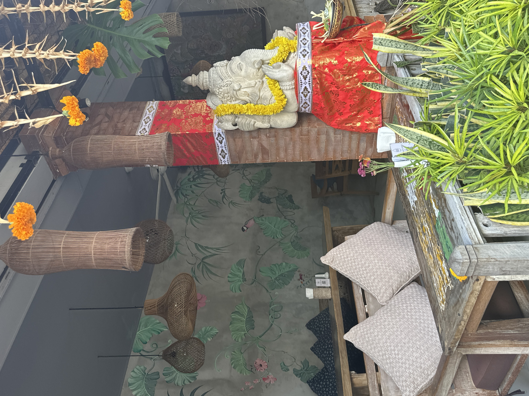 The image depicts an outdoor seating area, likely a restaurant or cafe, in Ubud District, Indonesia. A large stone statue of Ganesha, adorned with a yellow flower garland and draped in red fabric, is prominently featured on the right side of the frame. The statue stands beside a brick pillar wrapped in red cloth with gold patterns. The seating area includes wooden tables and benches with light-colored woven cushions. In the background, a wall is decorated with a mural of lush green foliage and a small bird. Woven pendant lamps hang from the ceiling. Several orange marigold flower garlands are suspended from the overhead structure, adding a touch of color and cultural decoration. The foreground is filled with various green plants, including snake plants. The overall ambiance suggests a tropical, culturally rich setting common in Bali.