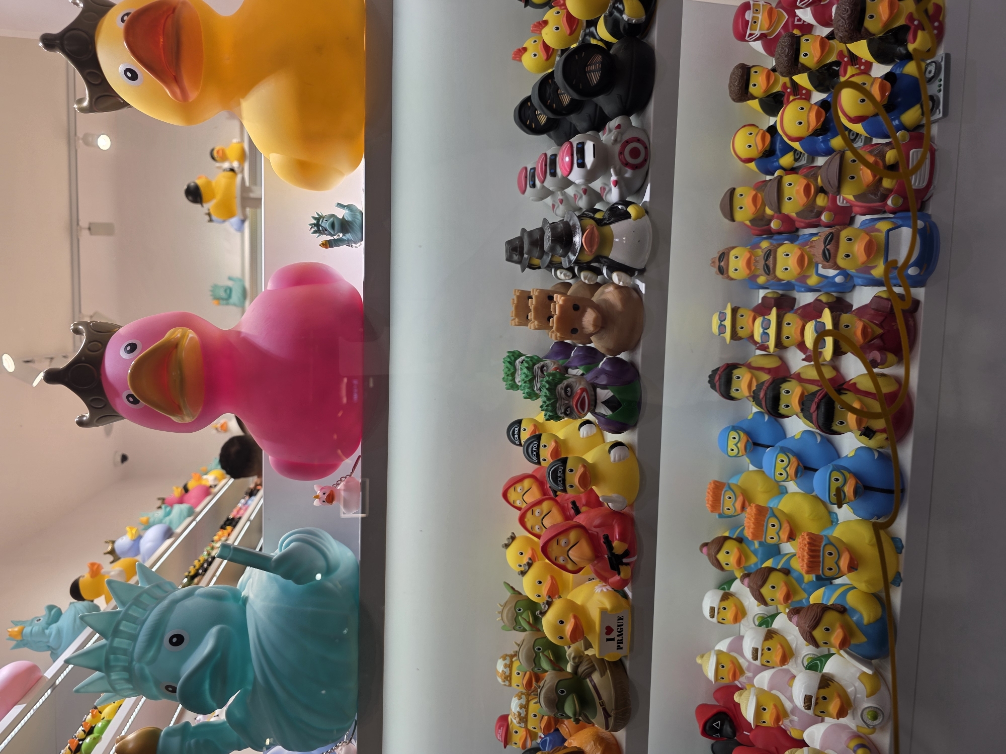The image displays a close-up view of a shelf filled with an assortment of rubber ducks. The ducks are arranged in rows and are all designed with various costumes and themes. In the foreground, several ducks are dressed as popular culture characters, including figures resembling the Joker, characters from the TV show "Money Heist" in red hooded outfits, and ducks dressed as robots and figures wearing hats.
Behind these, a larger collection of ducks is visible, many wearing hats, sunglasses, or themed outfits, such as a duck with an "I LOVE PRAGUE" sign. On the shelf above, larger, more stylized rubber ducks are displayed, including a teal-colored duck resembling the Statue of Liberty and a pink duck wearing a crown. To the right of these, a large yellow duck, also wearing a crown, is prominently featured. The background consists of a brightly lit display shelf, suggesting the ducks are for sale in a retail environment. No people are directly visible in the scene.