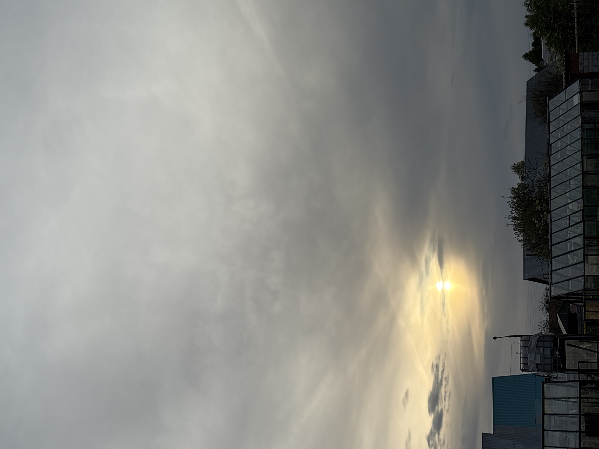 The image shows an outdoor scene in Прикамский, Russia, under an overcast sky with the sun breaking through the clouds. In the foreground and middle ground are various structures including what appear to be greenhouses or storage buildings with glass or plastic panels. One structure is blue. A streetlight is also visible. The sky dominates the upper portion of the image.