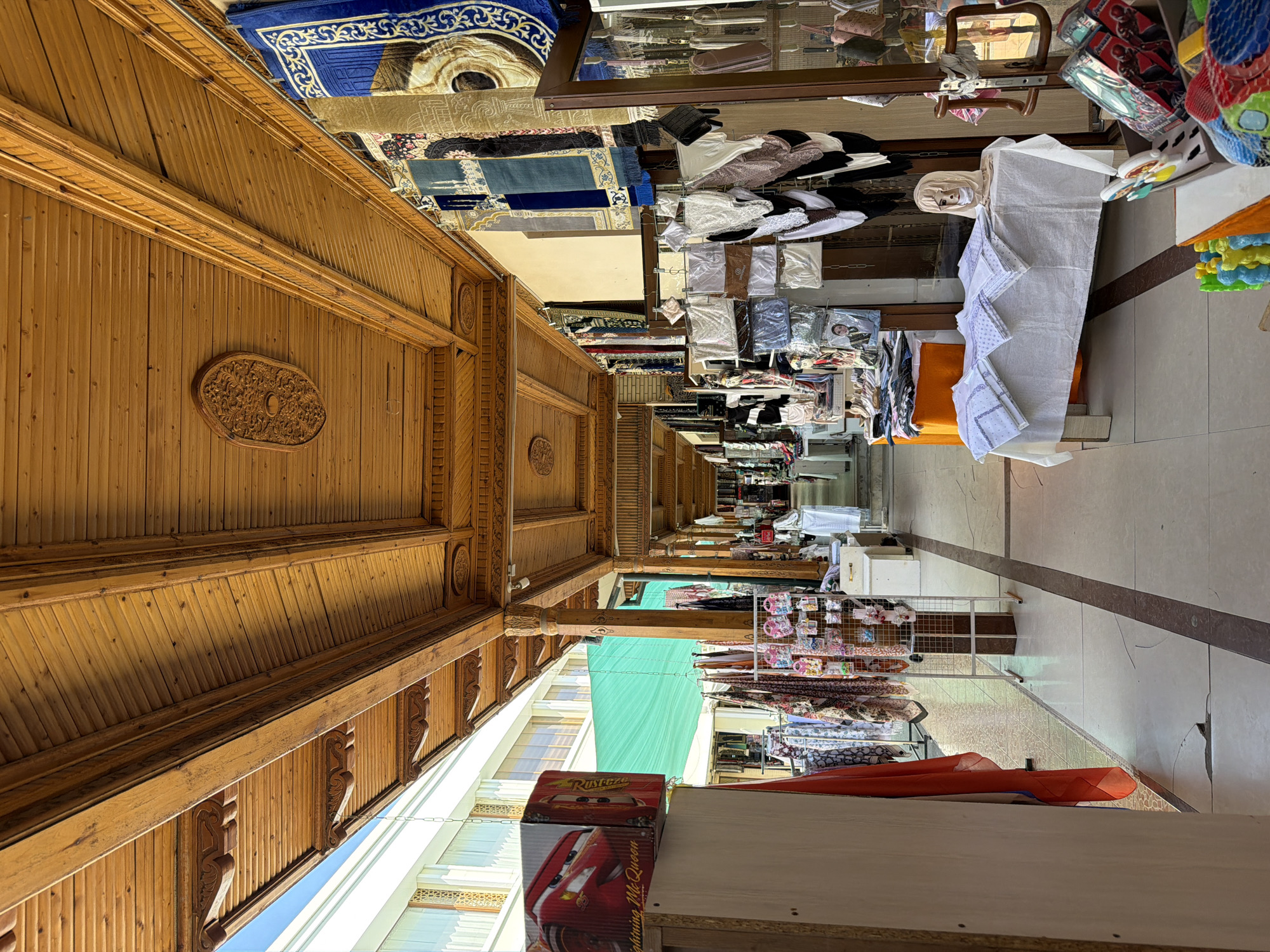The image captures an outdoor market with a covered walkway in Zangiata, Uzbekistan. The walkway is lined with stalls displaying various goods, including clothing, textiles, and small decorative items. Ornate wooden beams and intricate carvings adorn the ceiling of the covered area, suggesting traditional architecture. Sunlight filters through the open sides of the walkway, illuminating the colorful merchandise. In the foreground, a stall features a white tablecloth laden with folded patterned fabrics, and a mannequin head wearing a headscarf. Further down the walkway, more stalls are visible, with clothing racks and displays of scarves and other textiles. The floor is paved with tiles, and the overall atmosphere suggests a bustling marketplace.